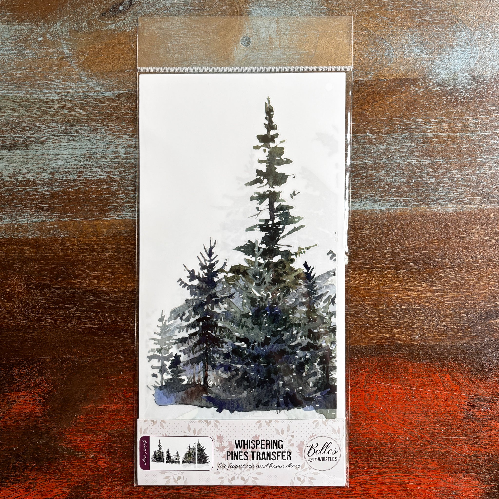 A package of Belles & Whistles' Whispering Pines rub-on transfer is against a dark wood background.