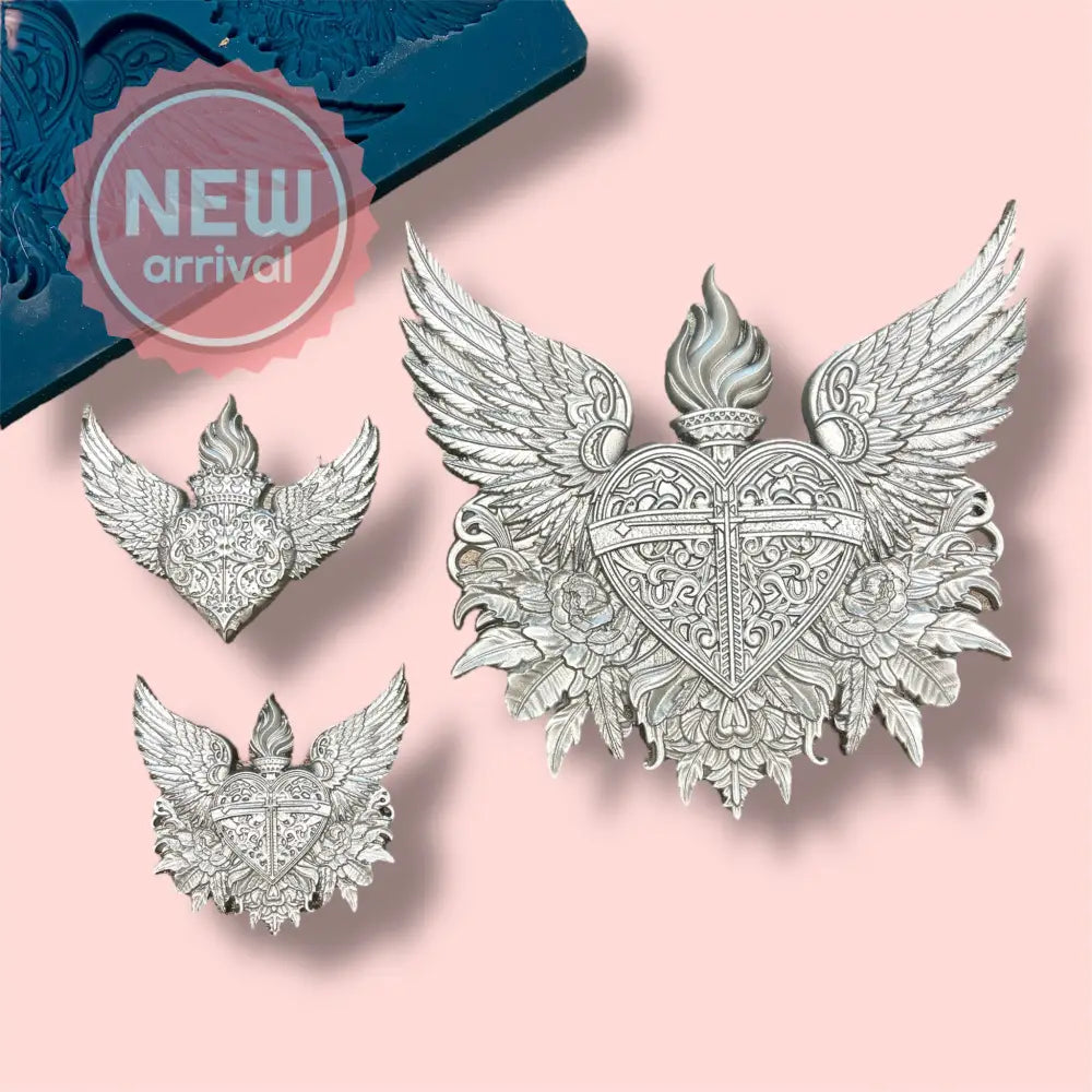 A blue silicone mold and silver colored castings featuring 2 small and 1 large heart, each topped with flames and surrounded by wings, all nestled upon delicate flowers are against a light pink background.