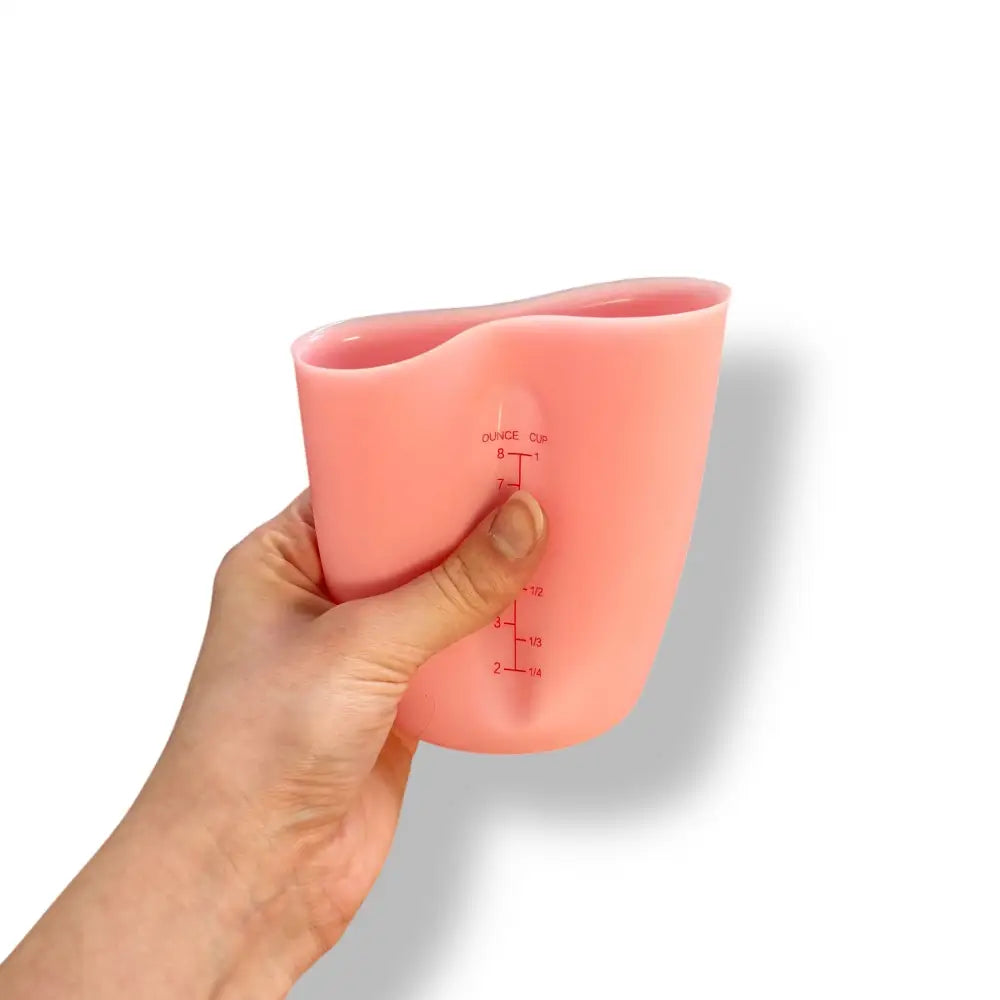 A hand is shown squeezing a light pink 8oz silicone measuring cup against a white background.