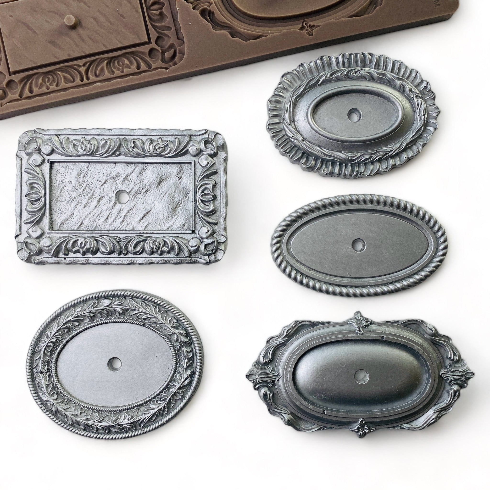 Silver colored silicone mold castings of 5 ornate drawer pull plates are against a white background.