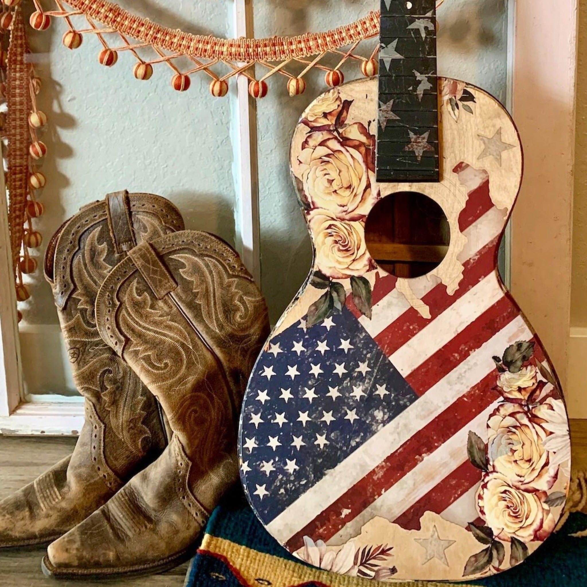 An acoustic guitar refurbished by Heartland Vintage Market features ReDesign with Prima's Sunflower Farms transfer on it along with an American Flag transfer. Cowboy boots sit to the left of the guitar.