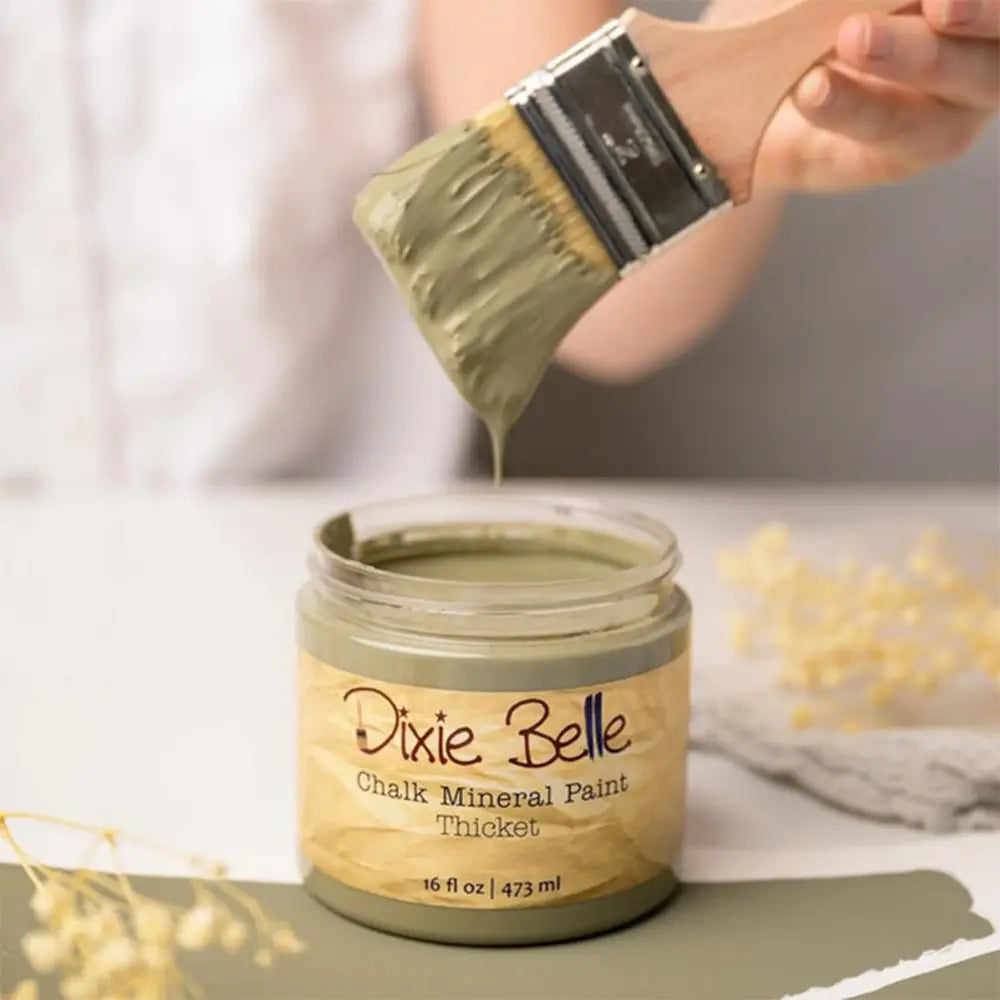 A paint brush with dripping paint is shown above an open container of Dixie Belle's Thicket Chalk Mineral Paint.