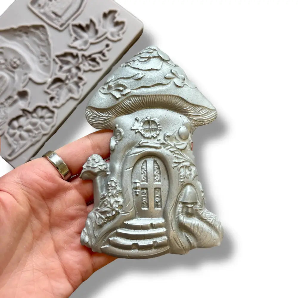 A light grey silicone mold featuring a mushroom fairy house, a hobbit style door, and leaves and flowers is against a white background. A hand is shown holding a silver colored casting of the mushroom house.