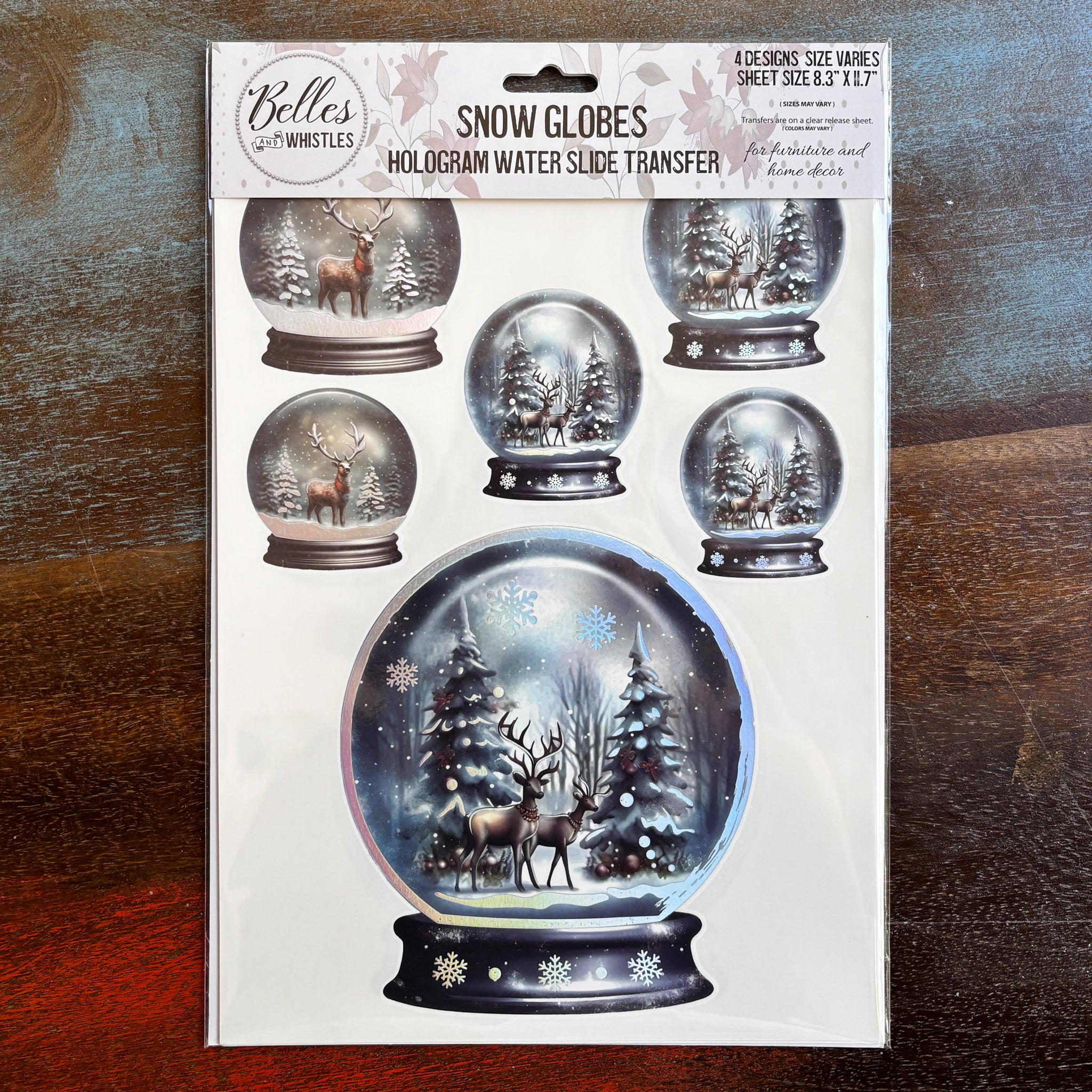 A package of Belles & Whistles' Snow Globes hologram water slide transfer is against a dark wood background.