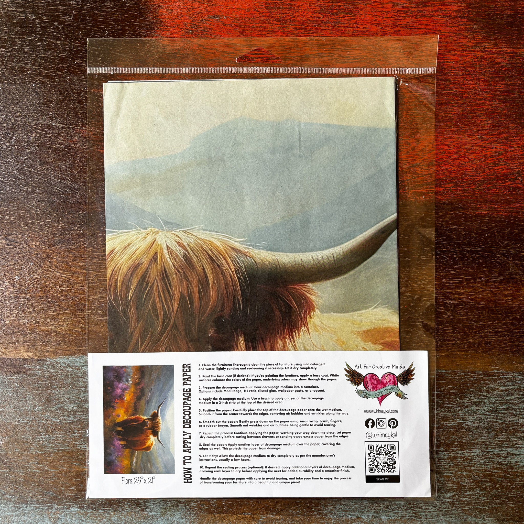 A package of Whimsykel's Floral Highland Cow tissue paper is against a dark wood background.