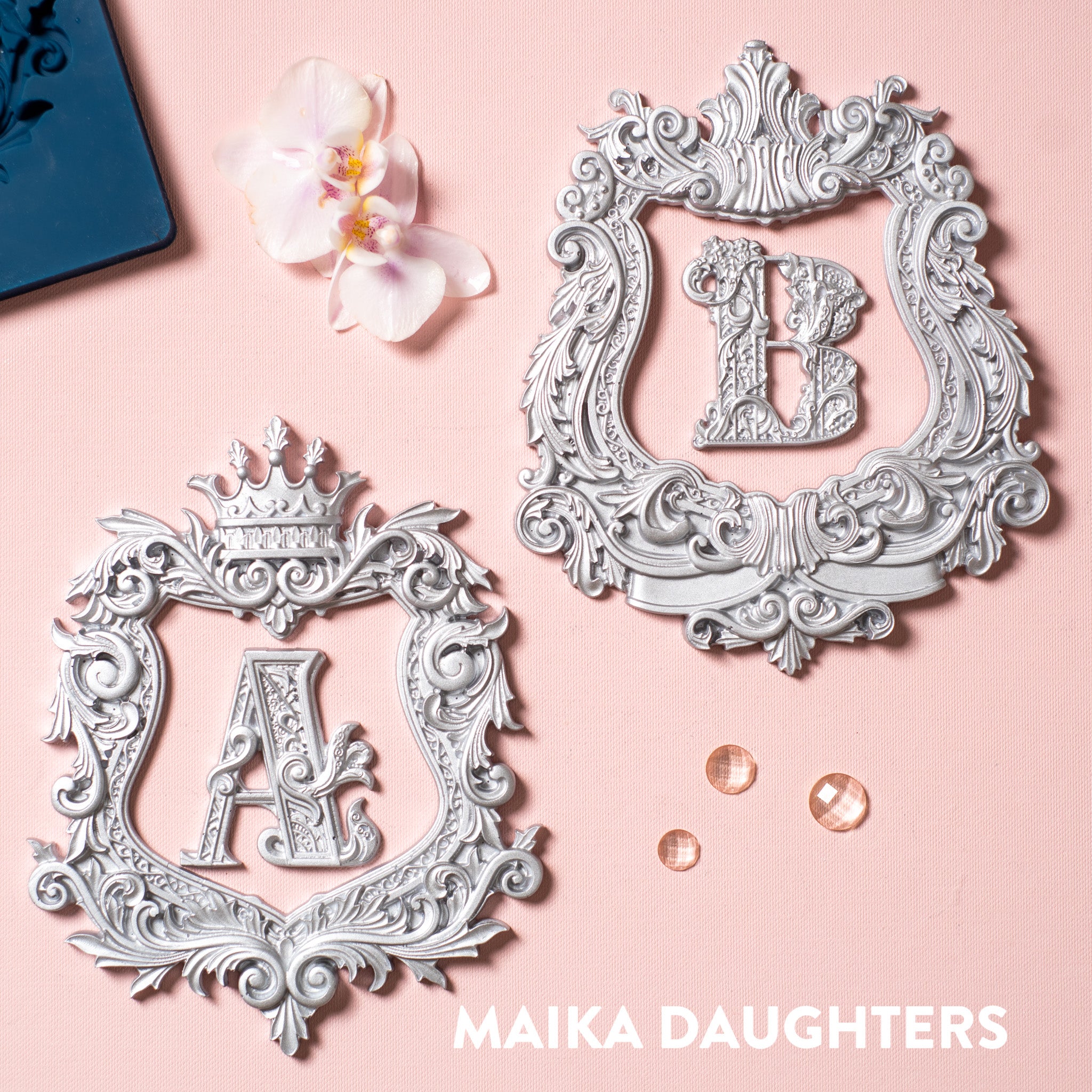 Silver colored castings made from a silicone mold that feature 2 ornate frames and 2 ornate letters A&B are against a light pink background.