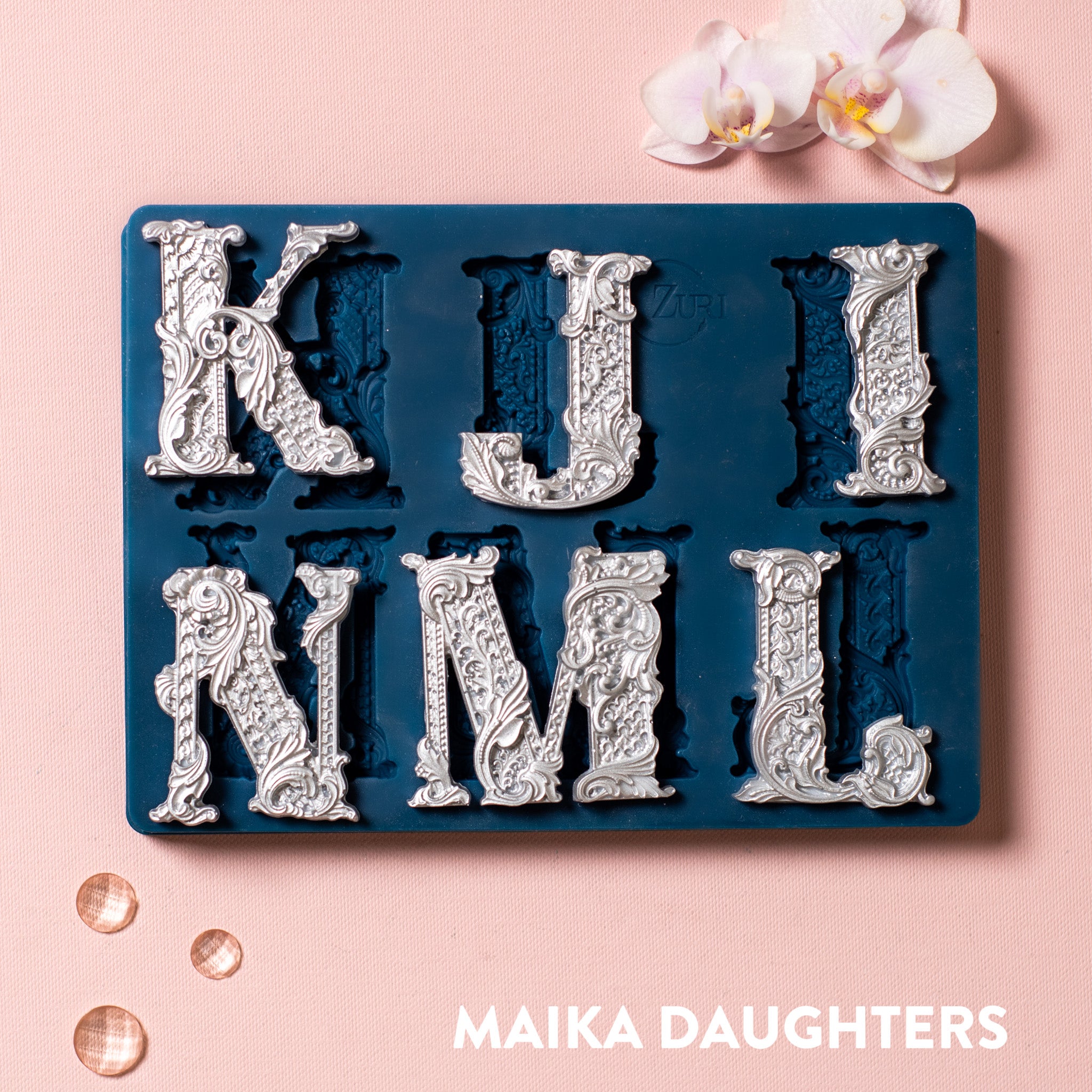 A blue silicone mold and silver colored castings of Zuri Design's IJKLMN Monograms are against a light pink background.