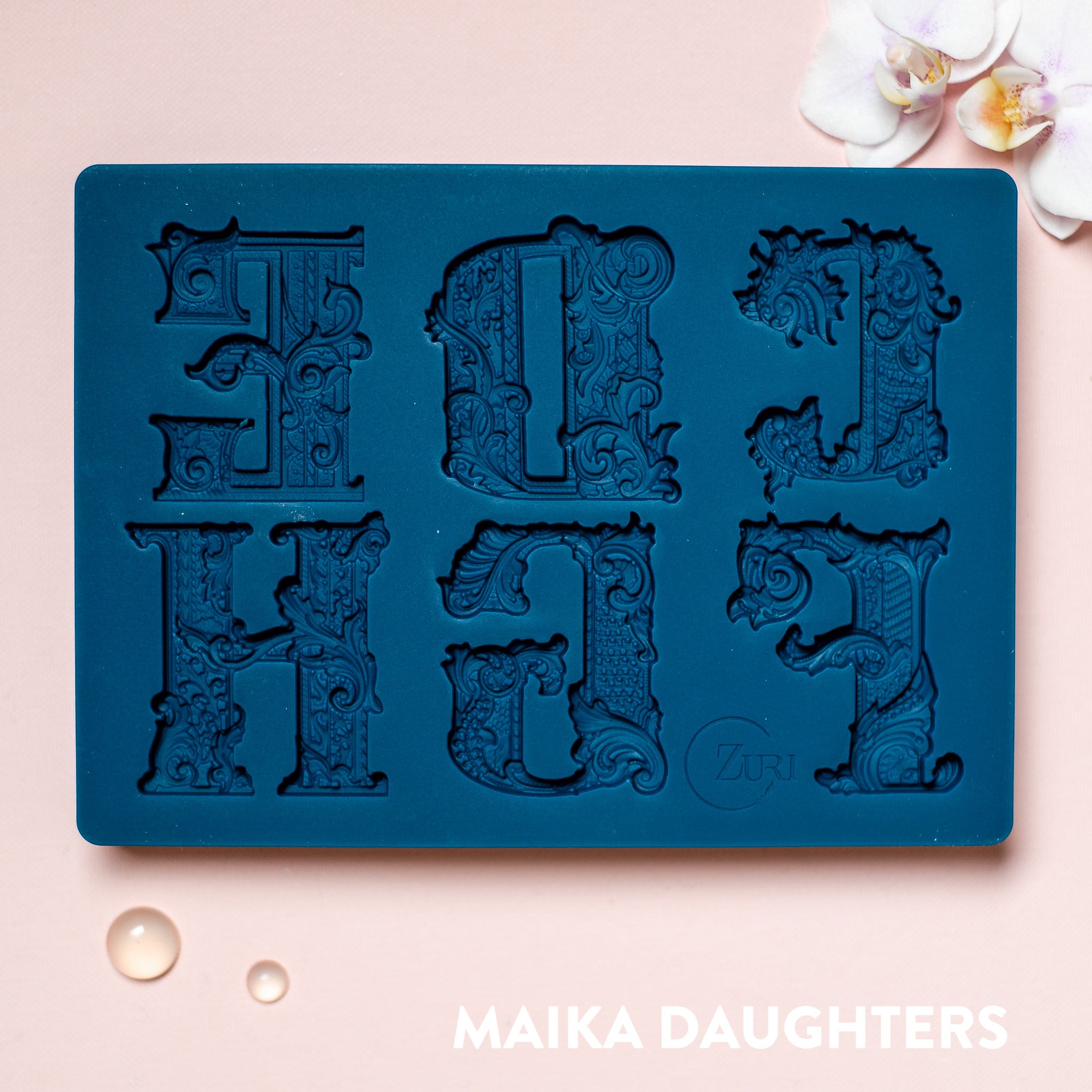 A blue silicone mold featuring 6 ornate letters of CDEFGH are against a light pink background.