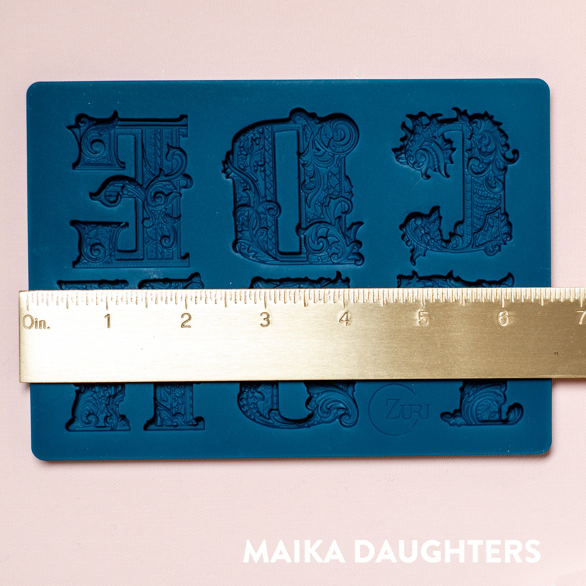 A blue silicone mold featuring 6 ornate letters of CDEFGH are against a light pink background. A gold ruler reading 6.73" width sits on the mold.