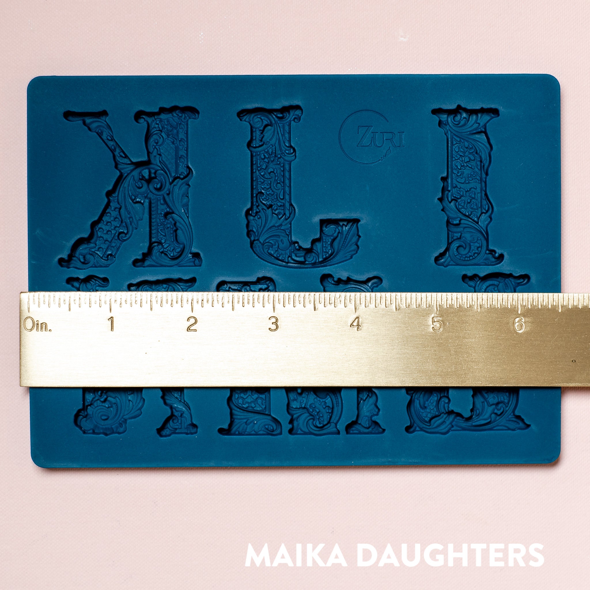 A blue silicone mold featuring 6 ornate letters of IJKLMN are against a light pink background. A gold ruler reading 6.73" width sits on the mold.