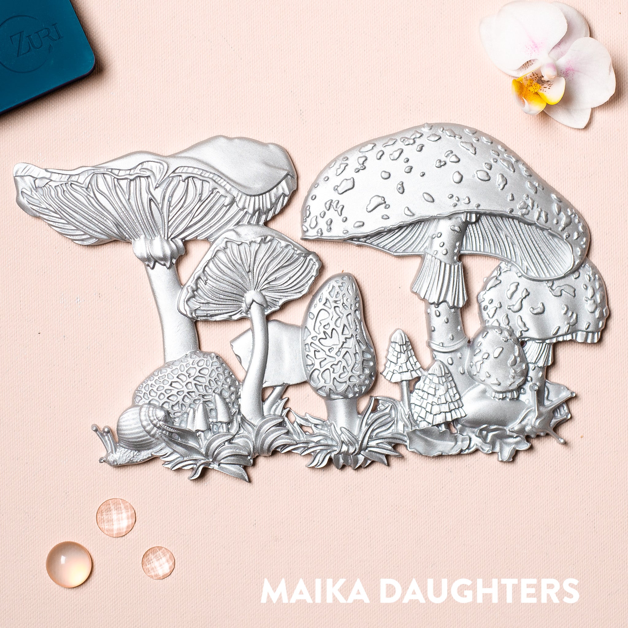 A silver casting of mushrooms against a pink background. 
Below in white lettering it reads Maika Daughters