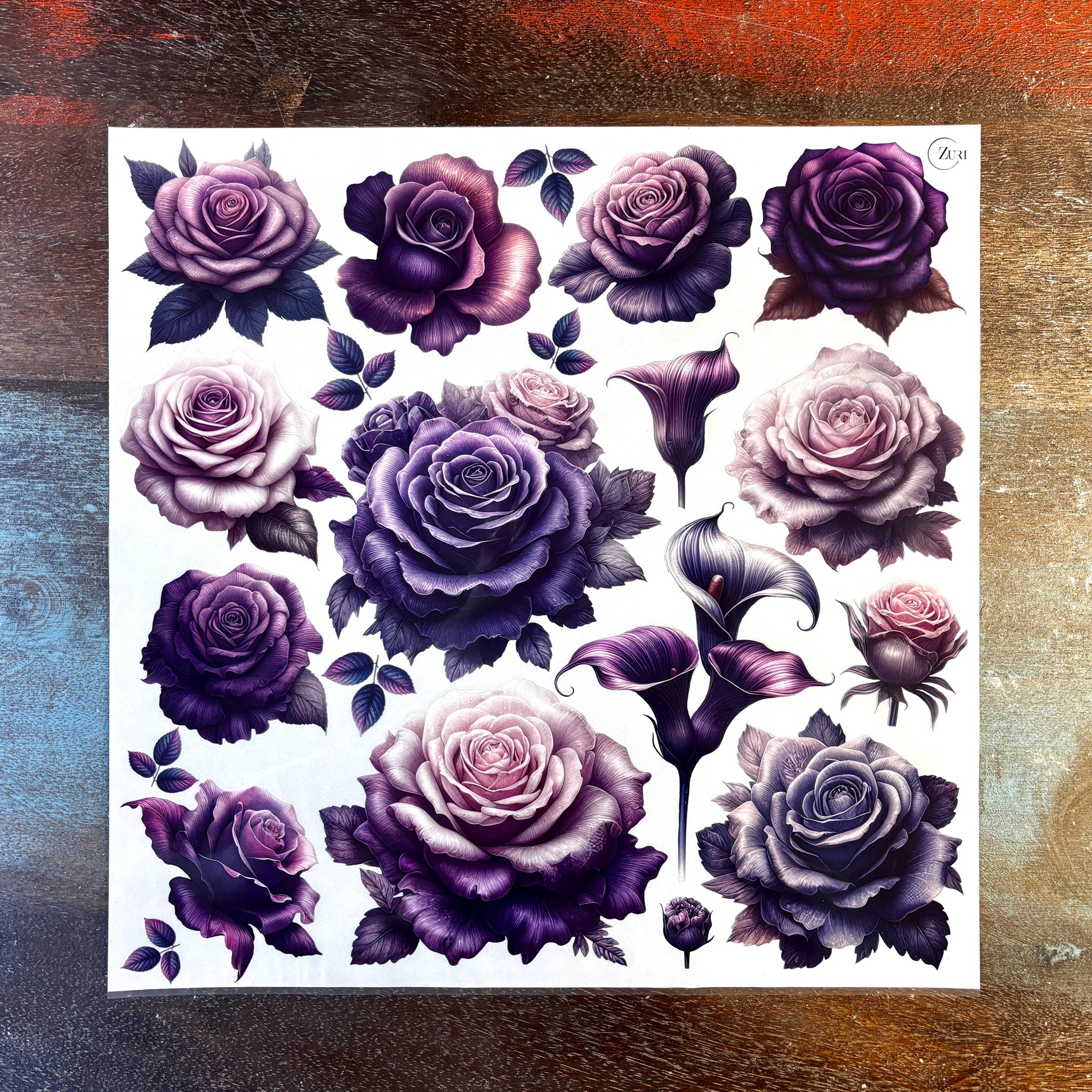 A sheet of small rub-on transfers featuring single blooms of roses and lilies in pinks and purples is against a dark wood background.