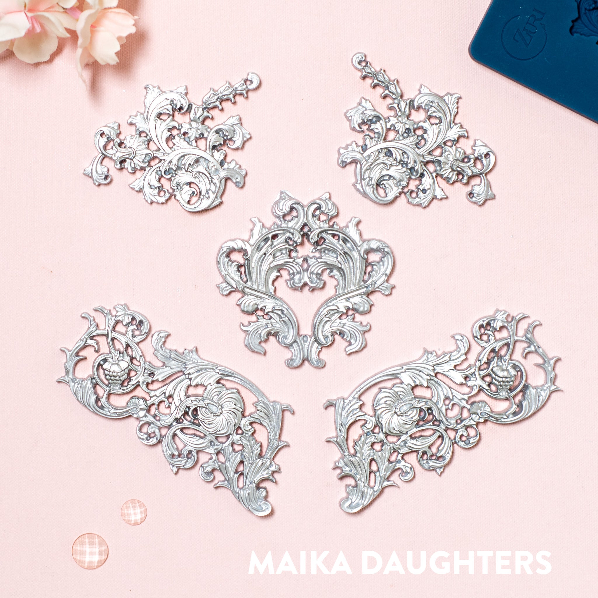 Silver colored silicone mold castings featuring 5 small ornate floral flourish accent pieces are against a pink background. Maika Daughters