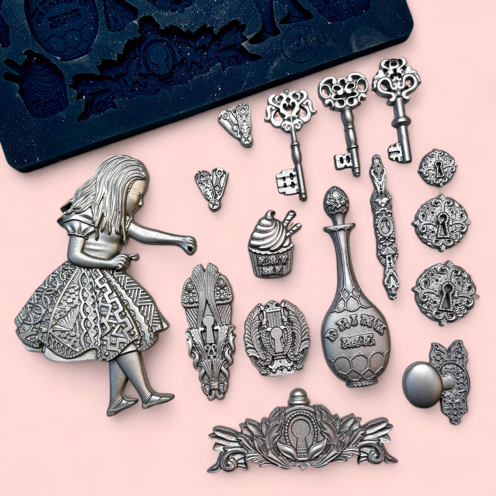 A blue silicone mold and silver colored silicone mold castings featuring Alice surrounded by keys, bottles, and other iconic images from Wonderland are against a light pink background.