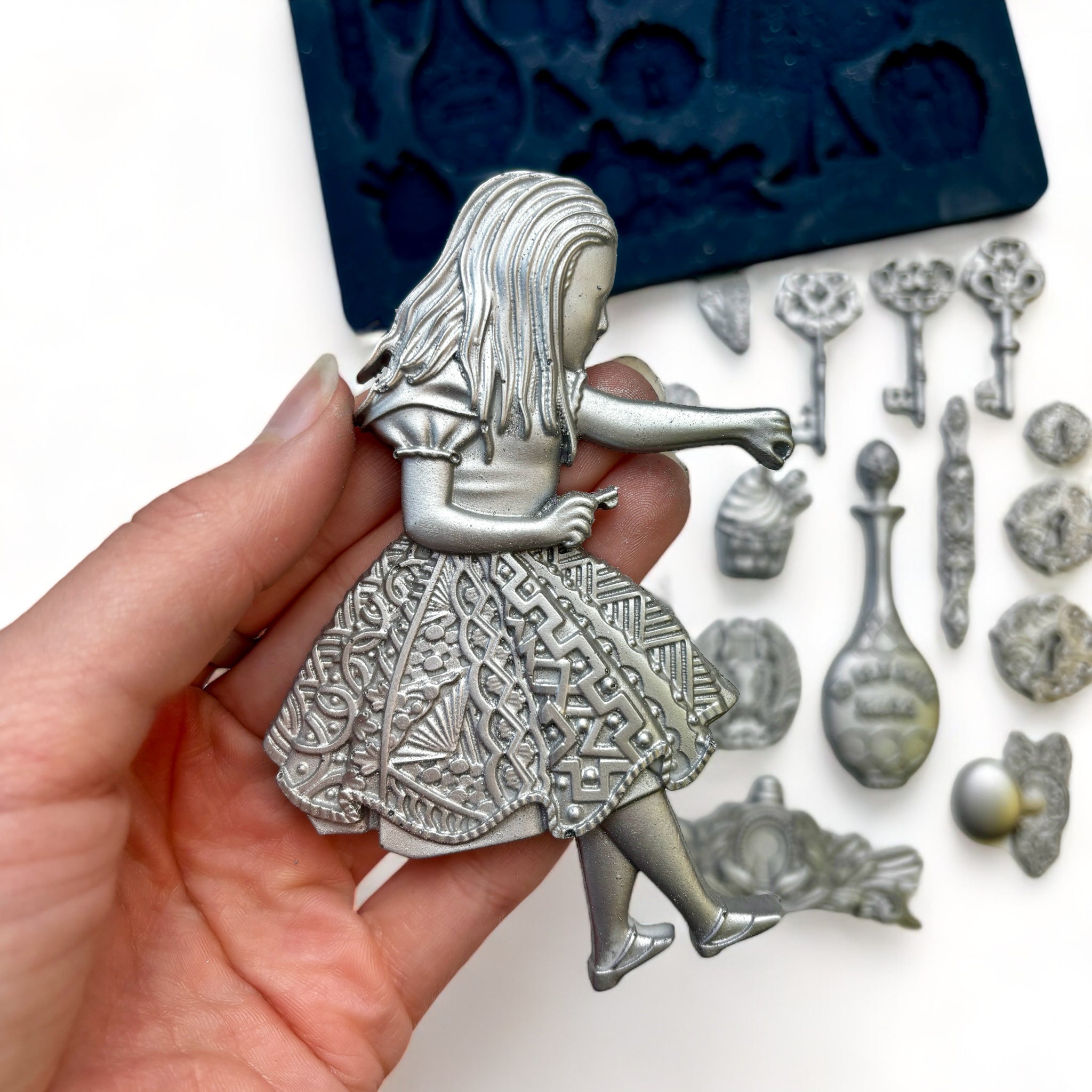 A blue silicone mold and silver colored castings of Zuri Design's Alice's Keys silicone mold are against a white background. A hand is shown holding the Alice casting.