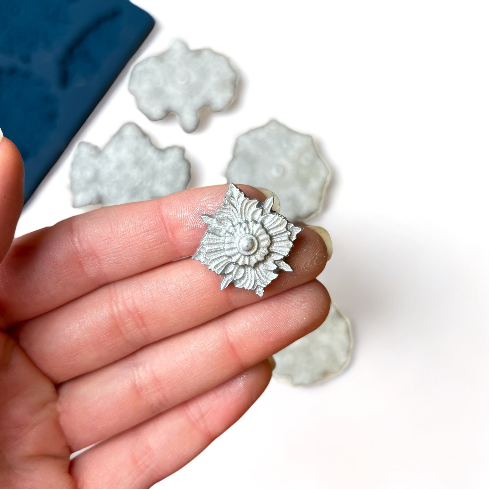 A blue silicone mold and silver colored castings of 10 varying size Baroque style medallion accents are against a white background. A hand is shown holding one of the smaller castings.