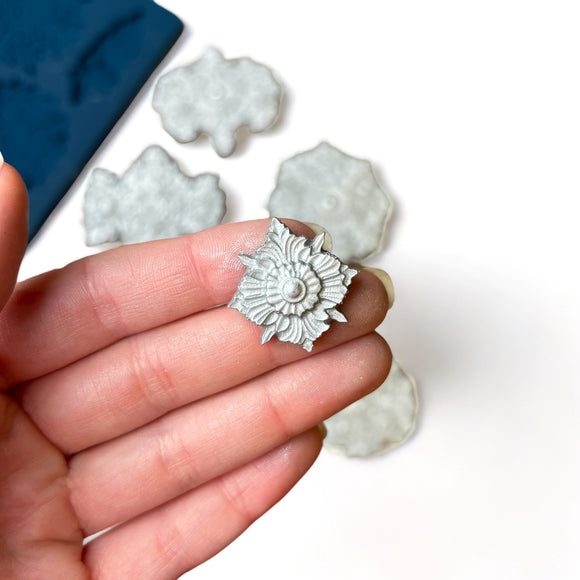 A blue silicone mold and silver colored castings of 10 varying size Baroque style medallion accents are against a white background. A hand is shown holding one of the smaller castings.