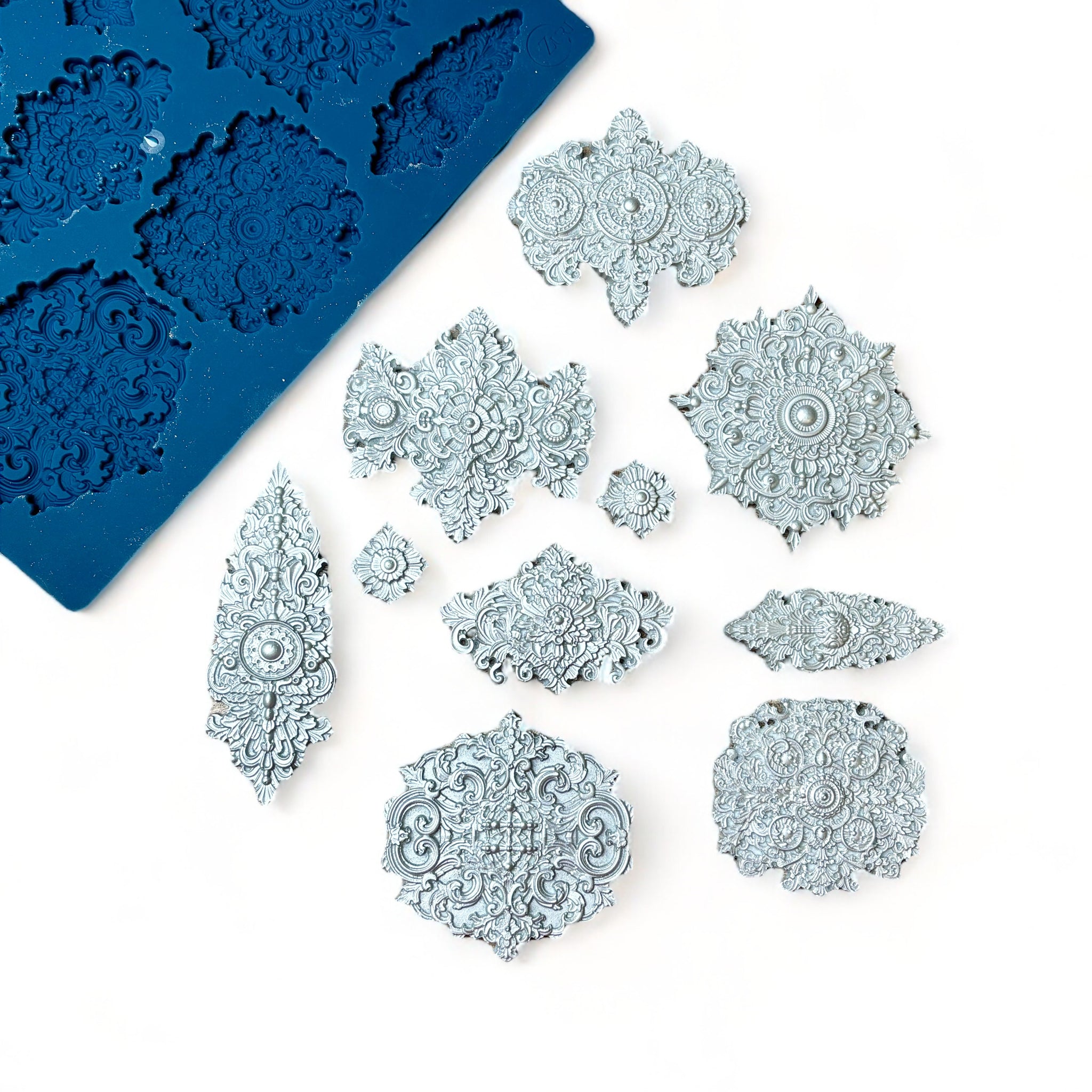 A blue silicone mold and silver colored castings of 10 varying size Baroque style medallion accents are against a light pink background.