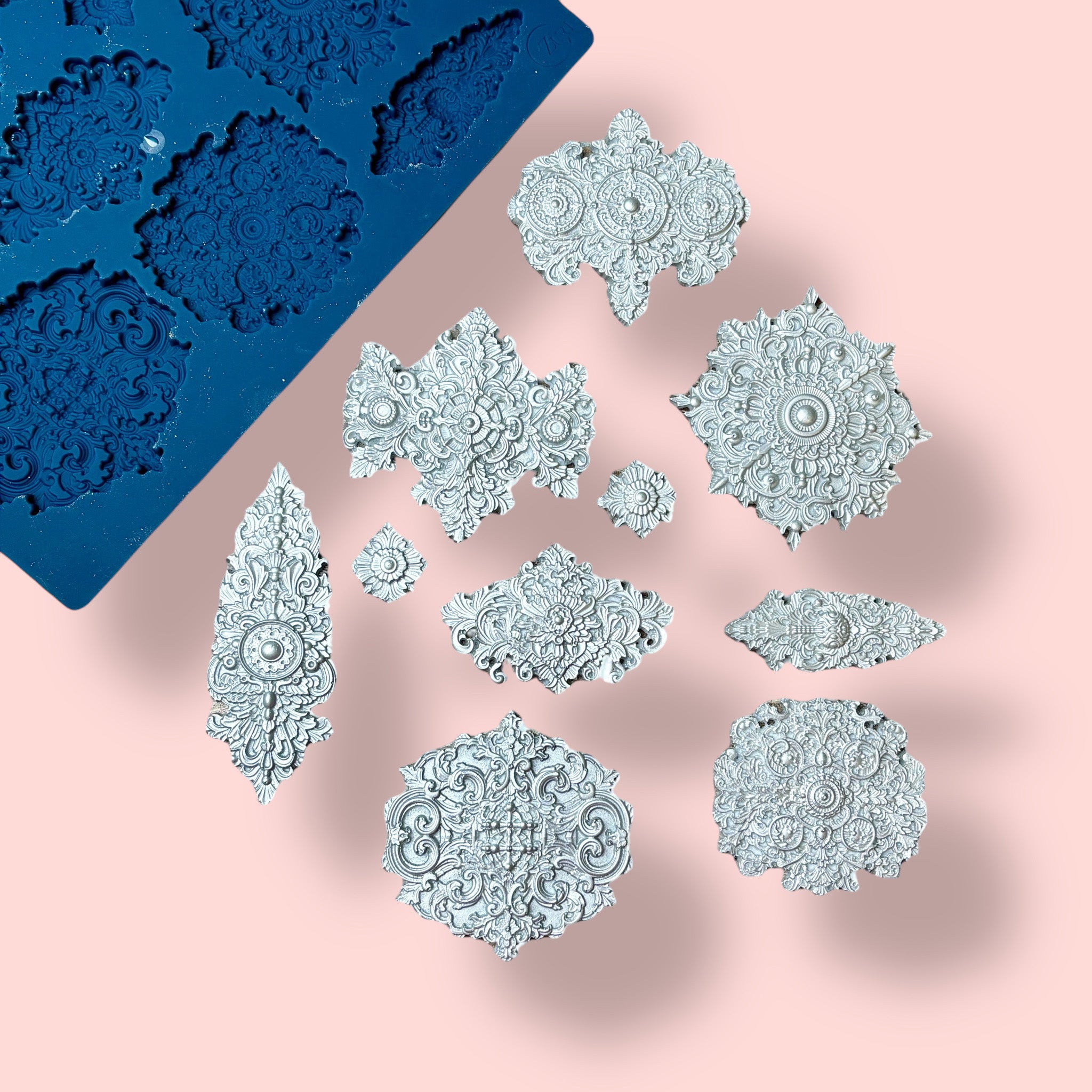 A blue silicone mold and silver colored castings of 10 varying size Baroque style medallion accents are against a light pink background.