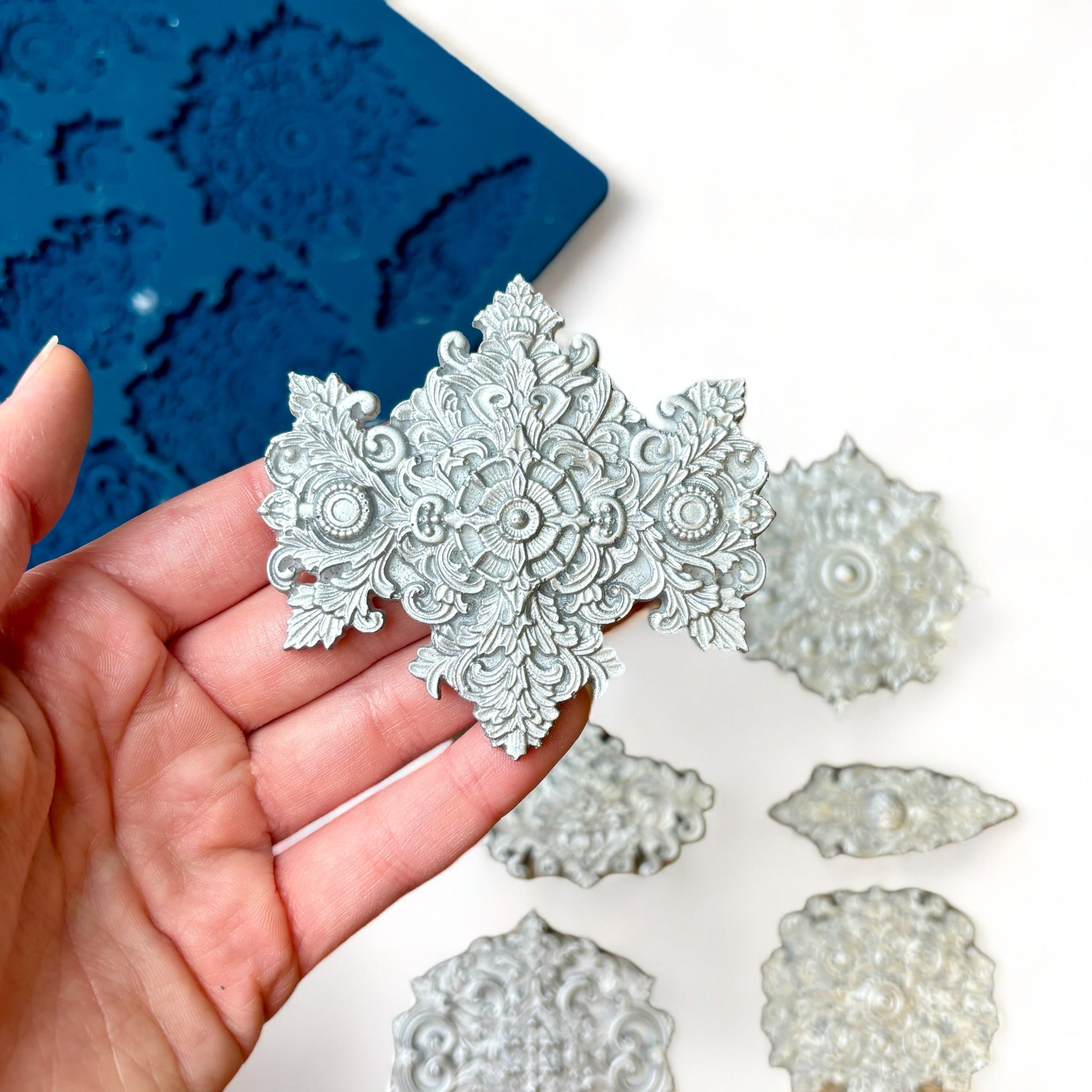 A blue silicone mold and silver colored castings of 10 varying size Baroque style medallion accents are against a white background. A hand is shown holding one of the larger castings.