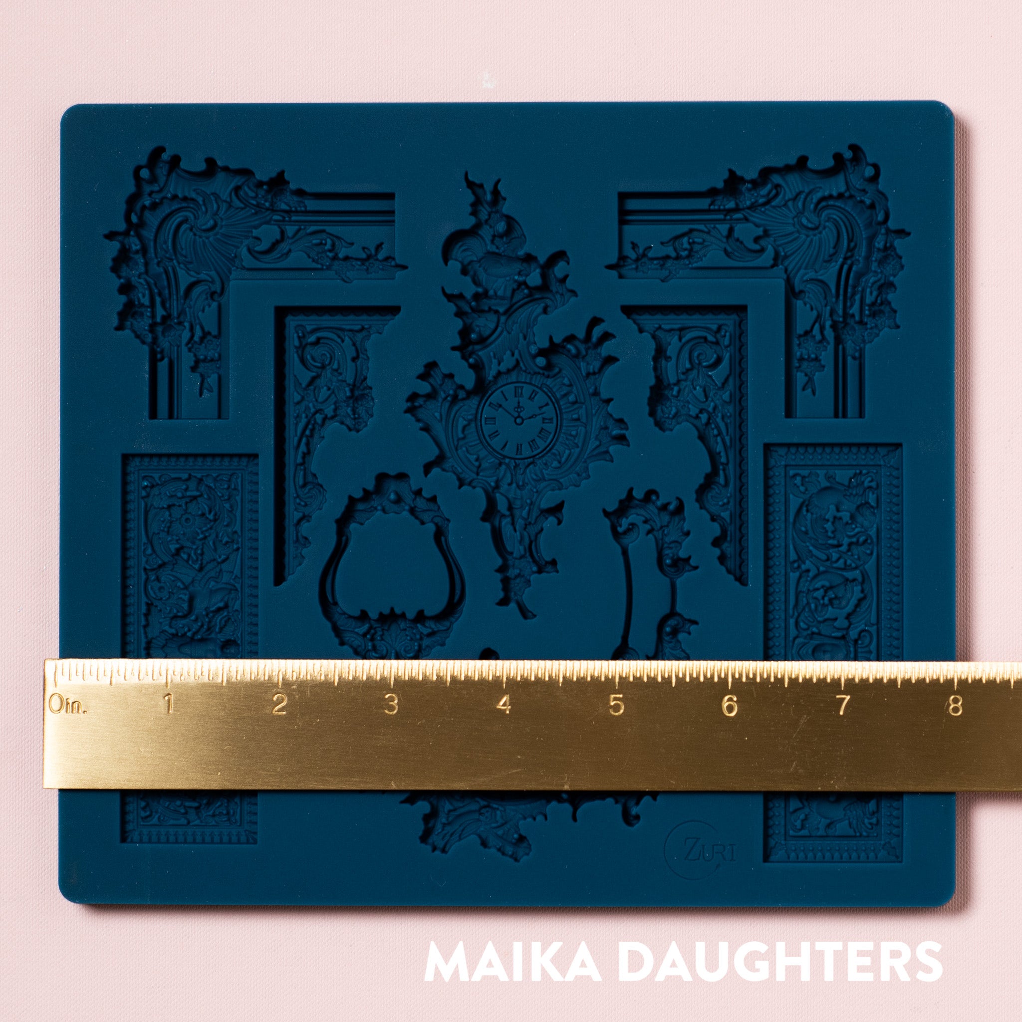 A blue silicone mold featuring 10 pieces of ornate frame corners, borders, and center accent pieces are against a light pink background. A gold ruler reading 8.8" wide sits on the mold.