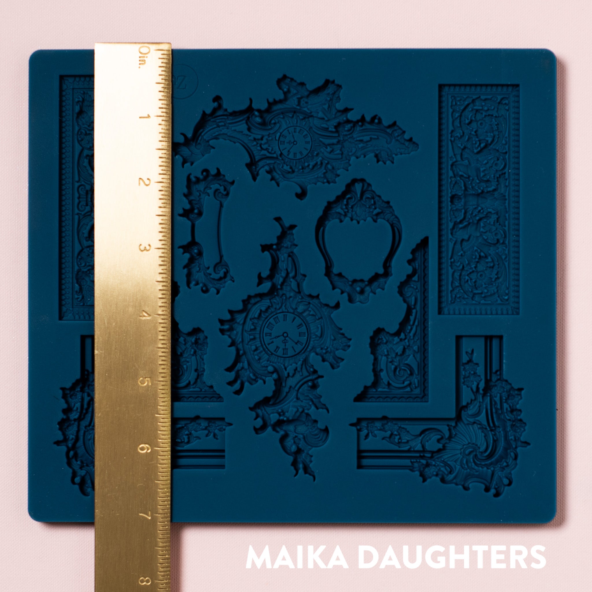 A blue silicone mold featuring 10 pieces of ornate frame corners, borders, and center accent pieces are against a light pink background. A gold ruler reading 5.4" height sits on the mold.