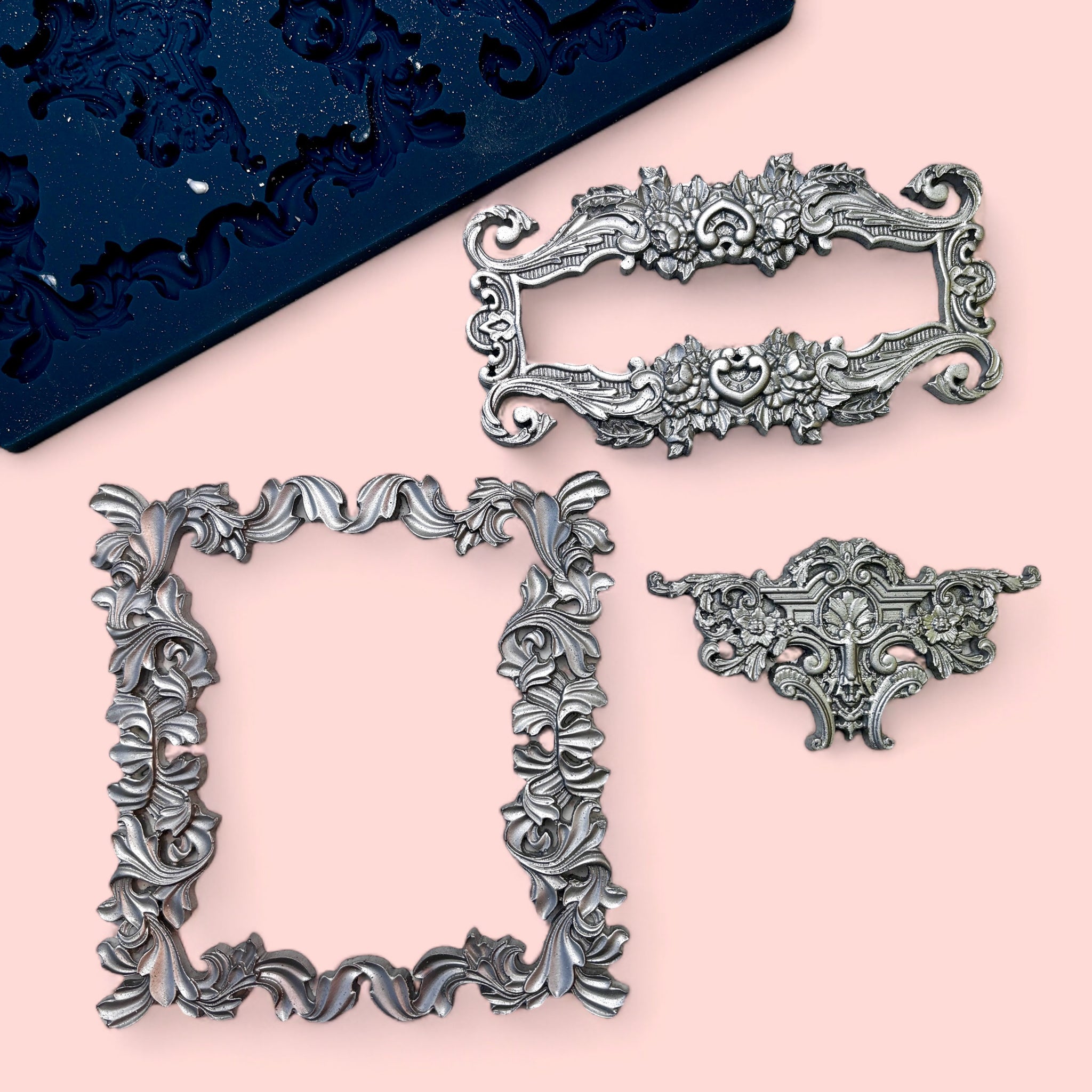 A blue silicone mold and silver colored casting of 2 ornate frames and a center accent piece are against a light pink background.