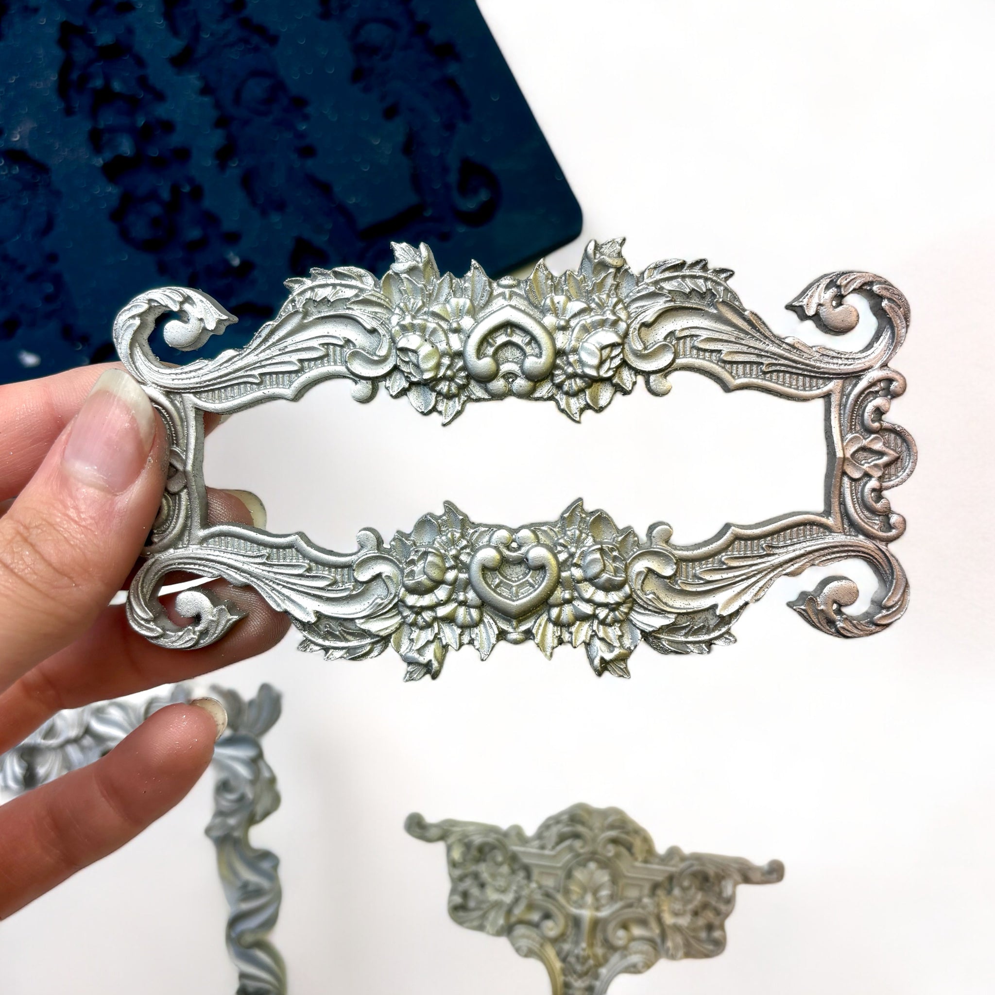 A blue silicone mold and silver colored castings of Zuri Design's Baroque Frames Set 2 are against a white background. A hand is shown holding one of the frames.