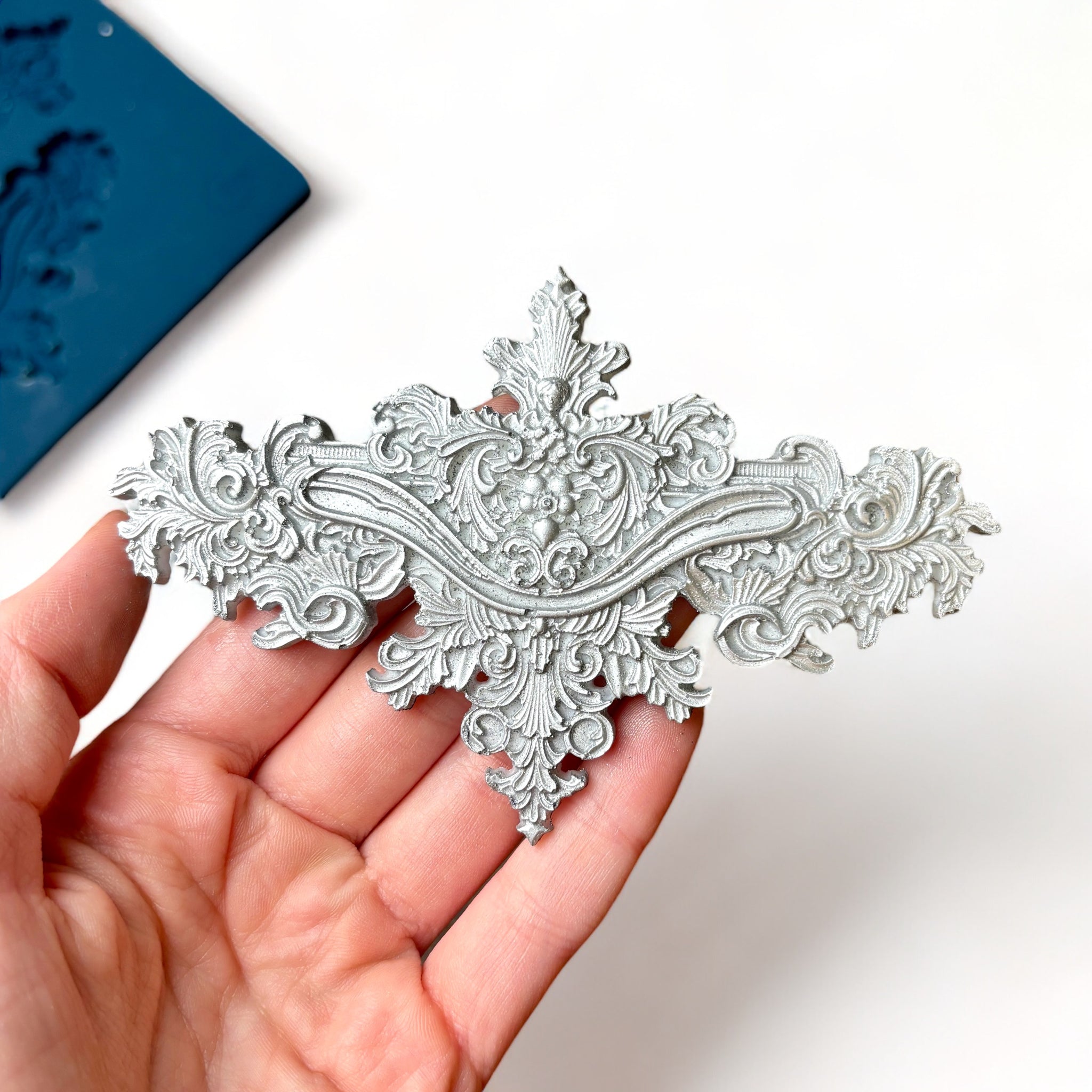 A blue silicone mold and silver colored castings of 2 large, and 2 small ornate baroque accent center pieces are against a white background. A hand is shown holding one of the large castings.