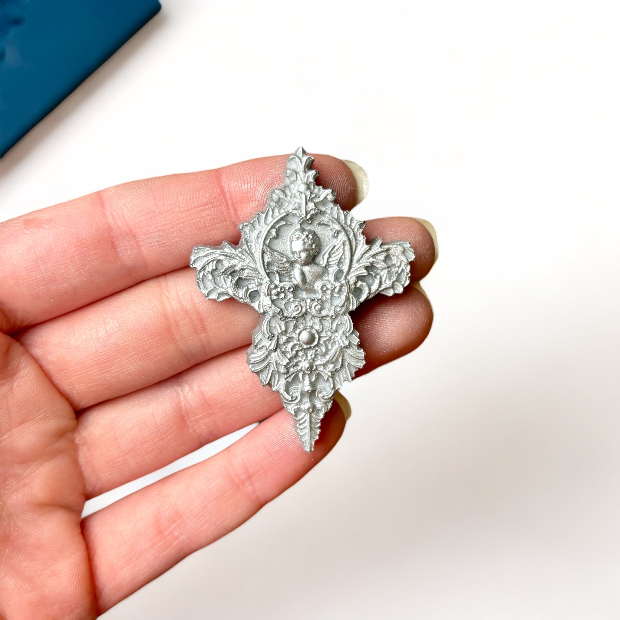 A blue silicone mold and silver colored castings of 2 large, and 2 small ornate baroque accent center pieces are against a white background. A hand is shown holding one of the small castings.