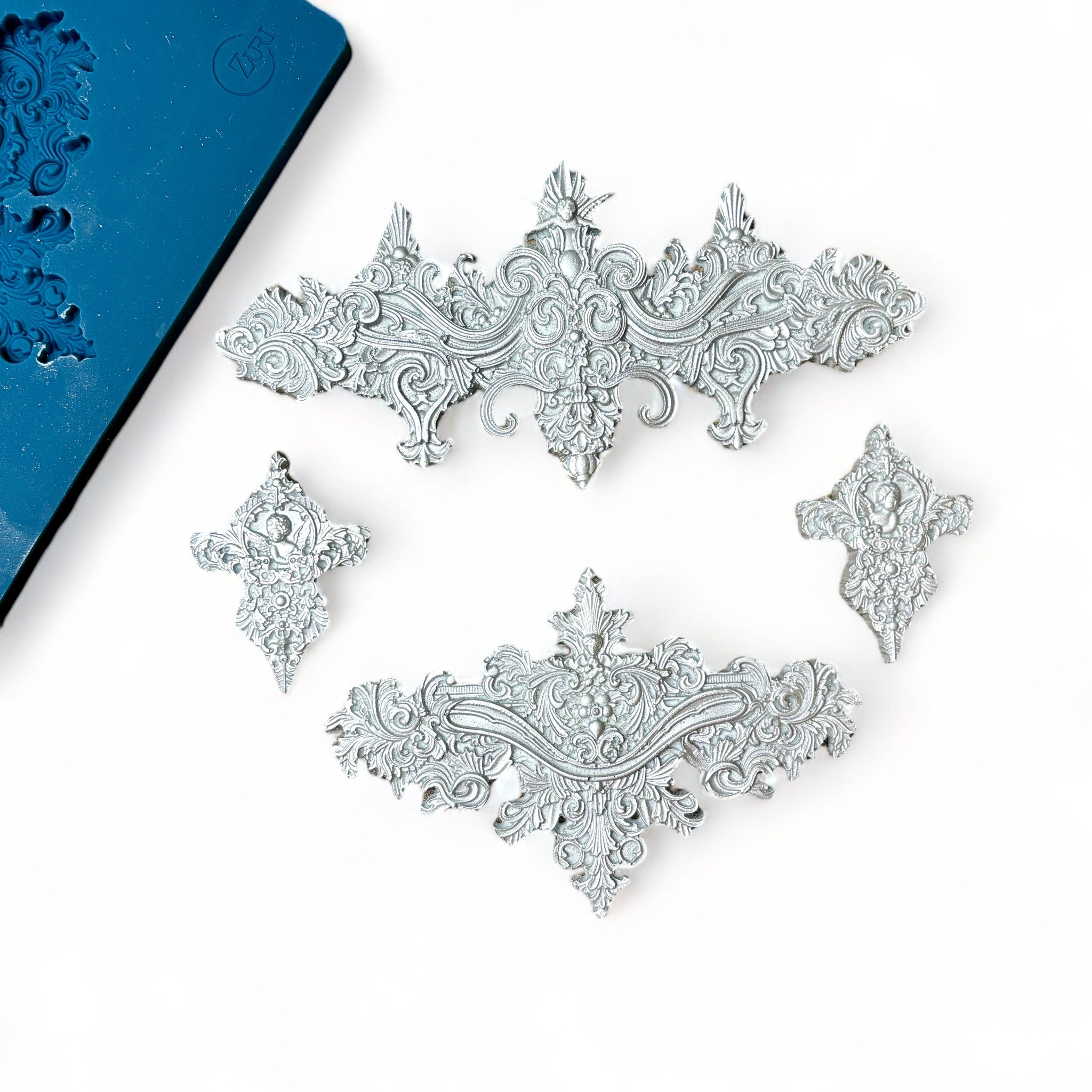 A blue silicone mold and silver colored castings of 2 large, and 2 small ornate baroque accent center pieces are against a light pink background.