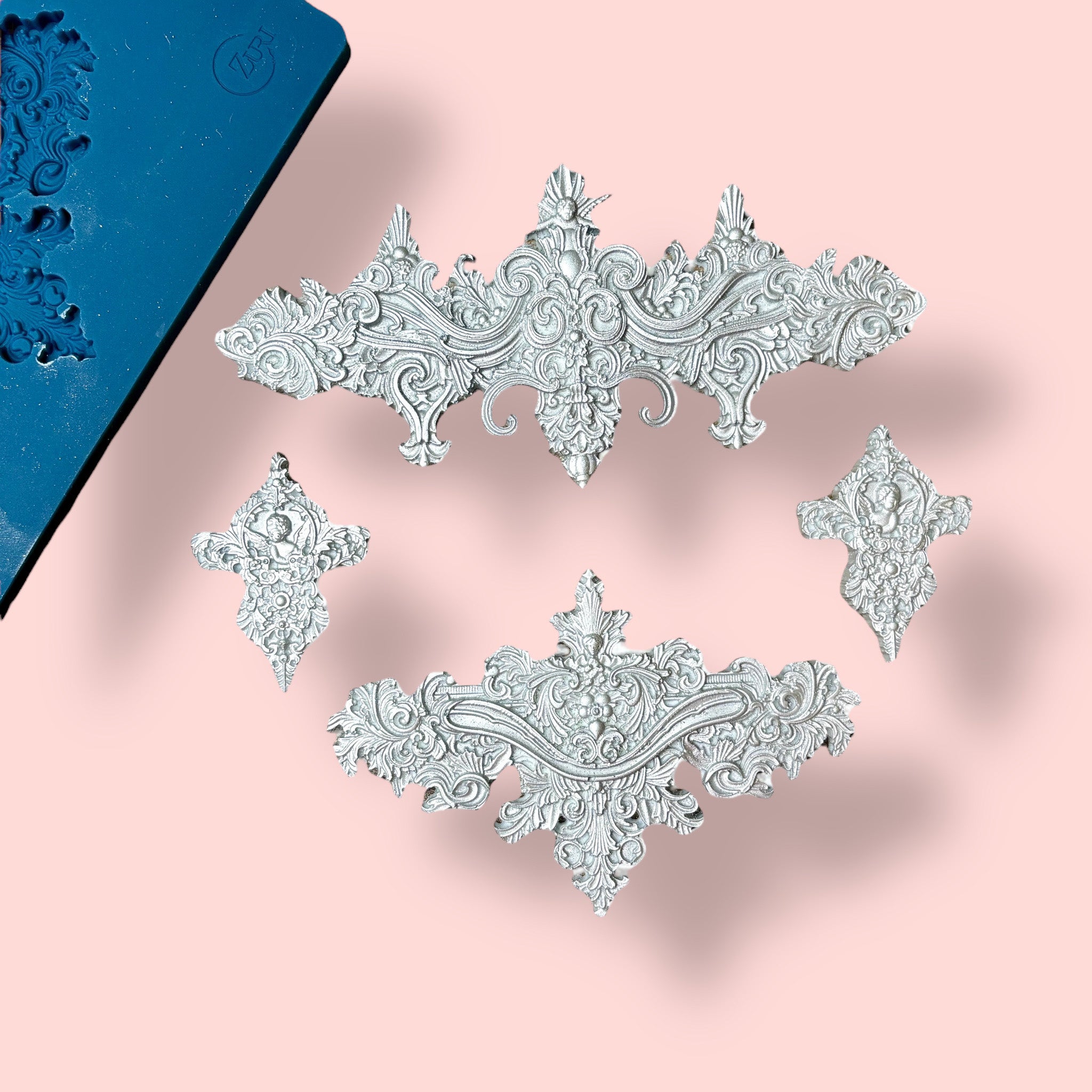 A blue silicone mold and silver colored castings of 2 large, and 2 small ornate baroque accent center pieces are against a light pink background.