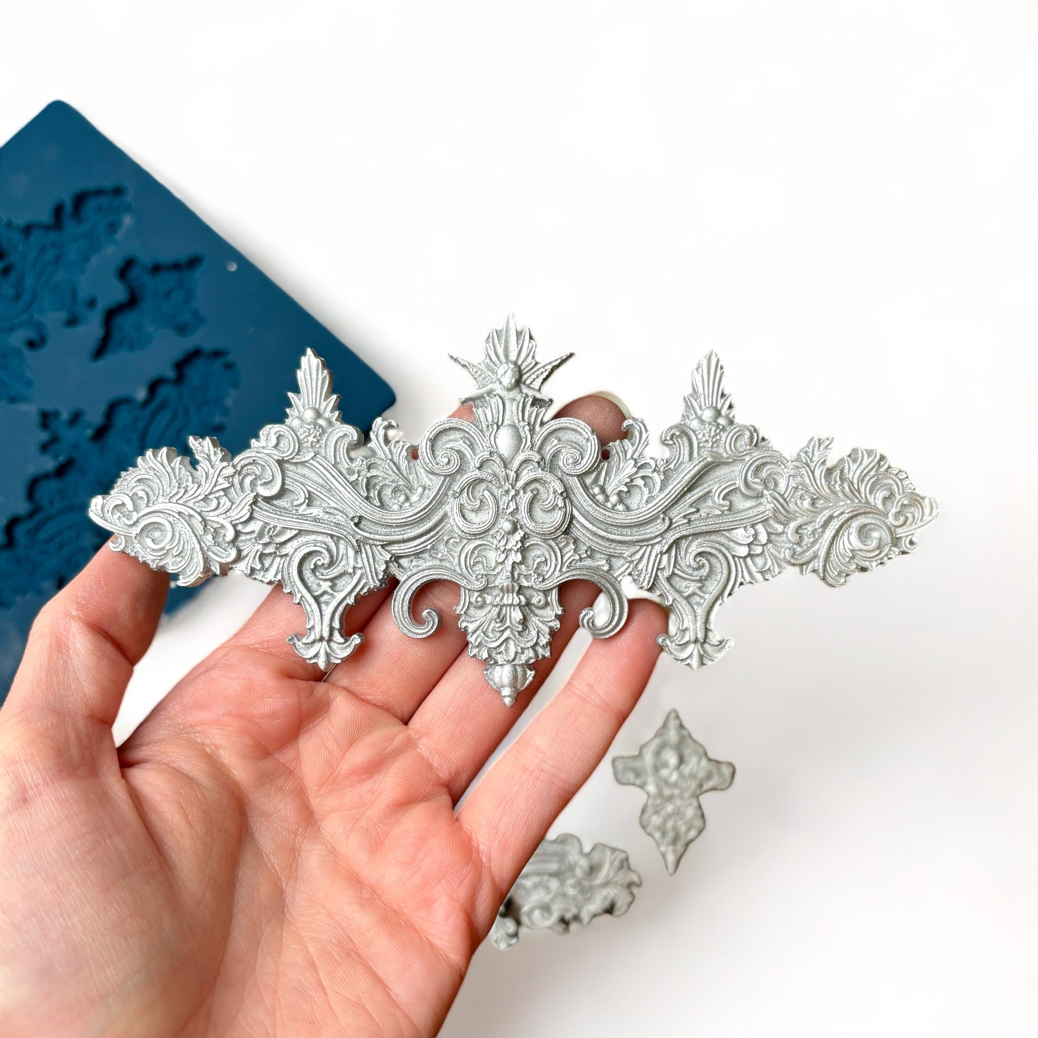 A blue silicone mold and silver colored castings of 2 large, and 2 small ornate baroque accent center pieces are against a white background. A hand is shown holding one of the large castings.