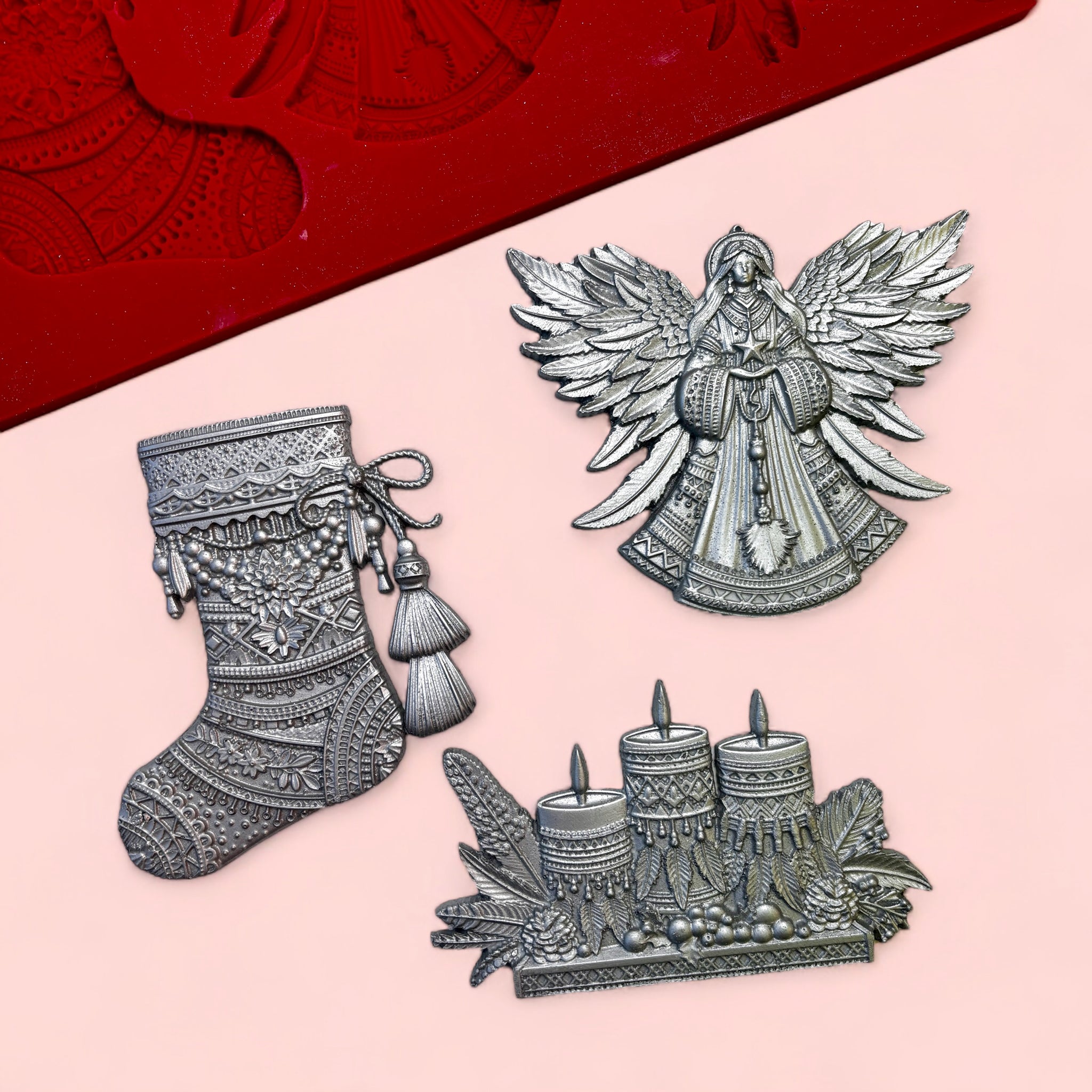 A red silicone mold and silver colored castings of a Bohemian style stocking, set of 3 candles and a Christmas angel are against a light pink background.