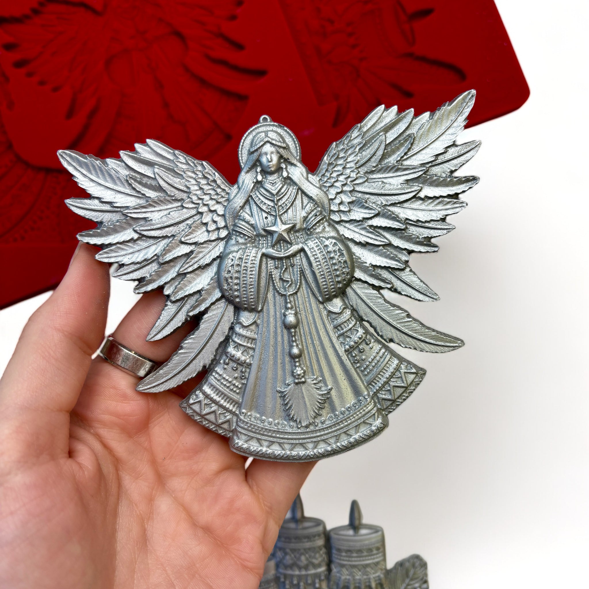 A red silicone mold and silver colored castings of Zuri Design's Bohemian Christmas Decor are against a white background. A hand is shown holding the angel casting.