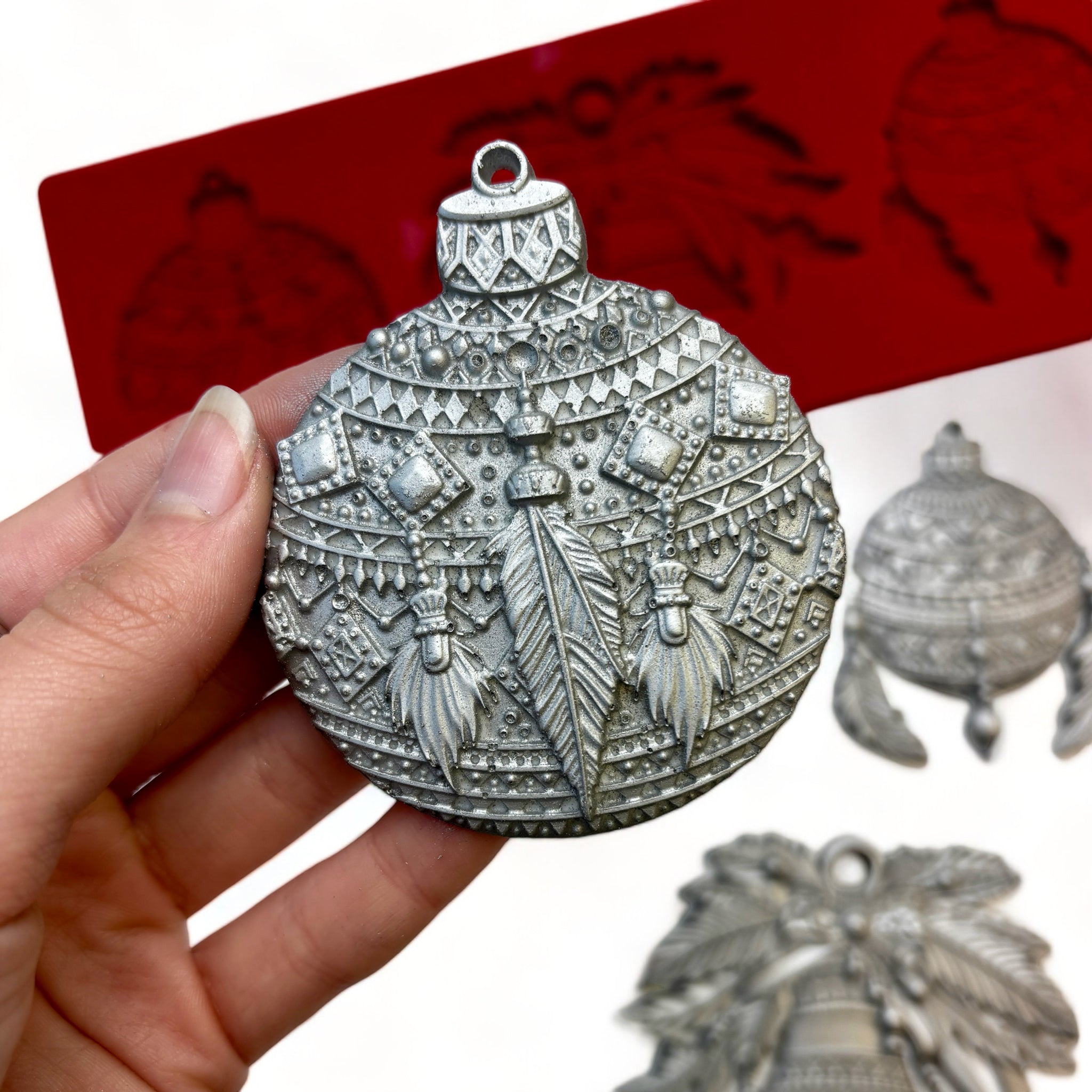 A red silicone mold and silver colored castings of Zuri Design's Bohemian Christmas Ornaments are against a white background. A hand is shown holding one of the ornaments.