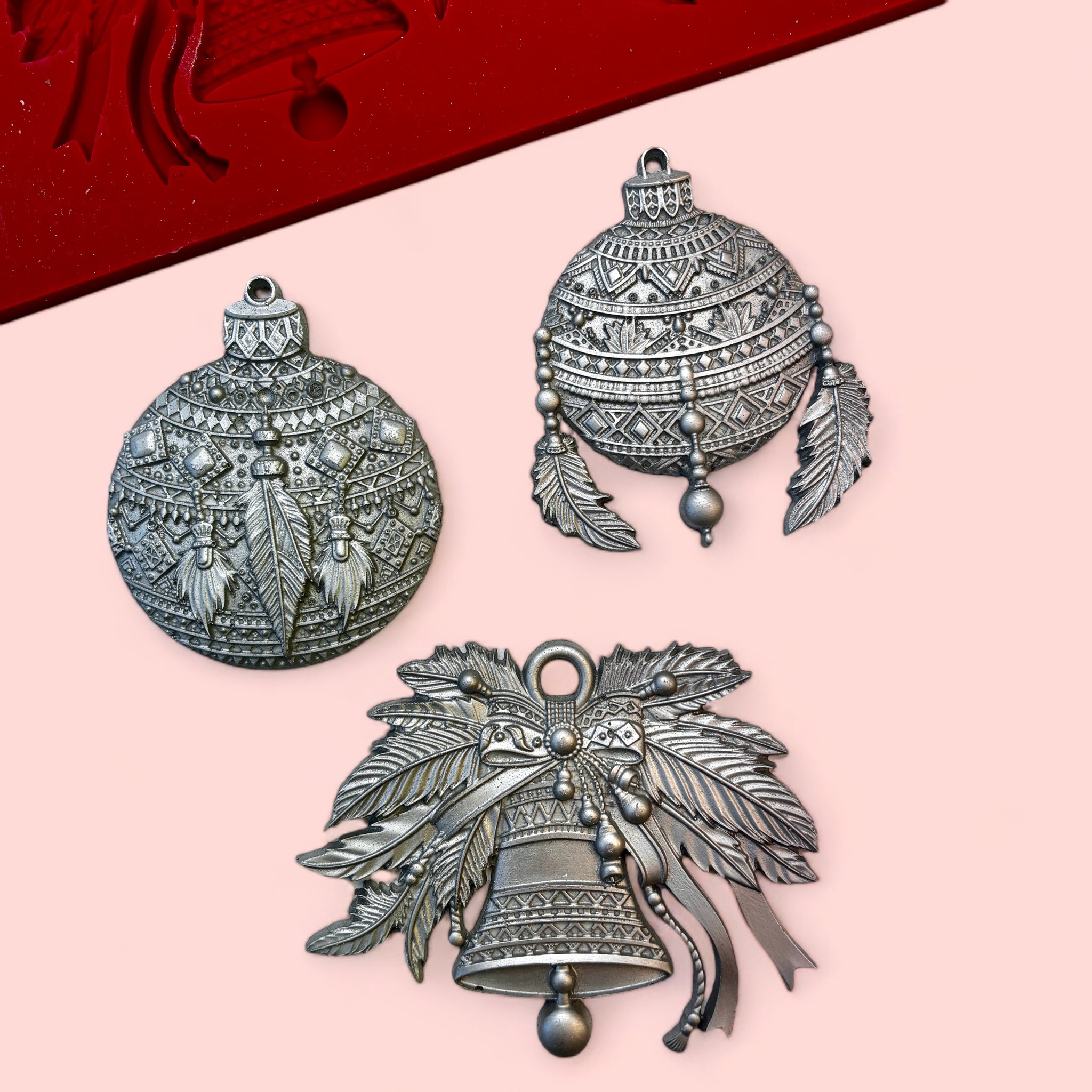 A red silicone mold and silver colored castings of 3 bohemian style Christmas ornaments are against a light pink background.