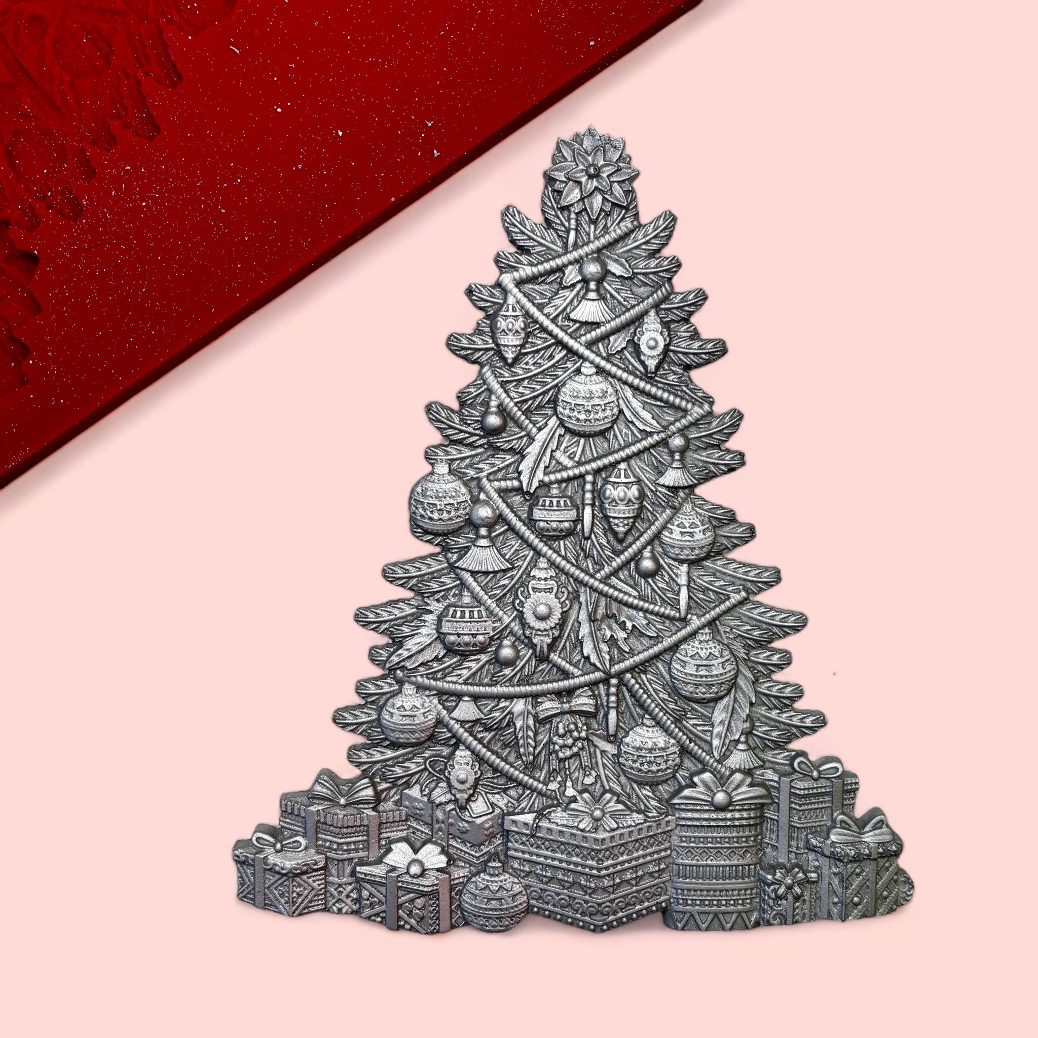 A red silicone mold and silver colored casting of a Bohemian style decorated Christmas tree are against a light pink background.