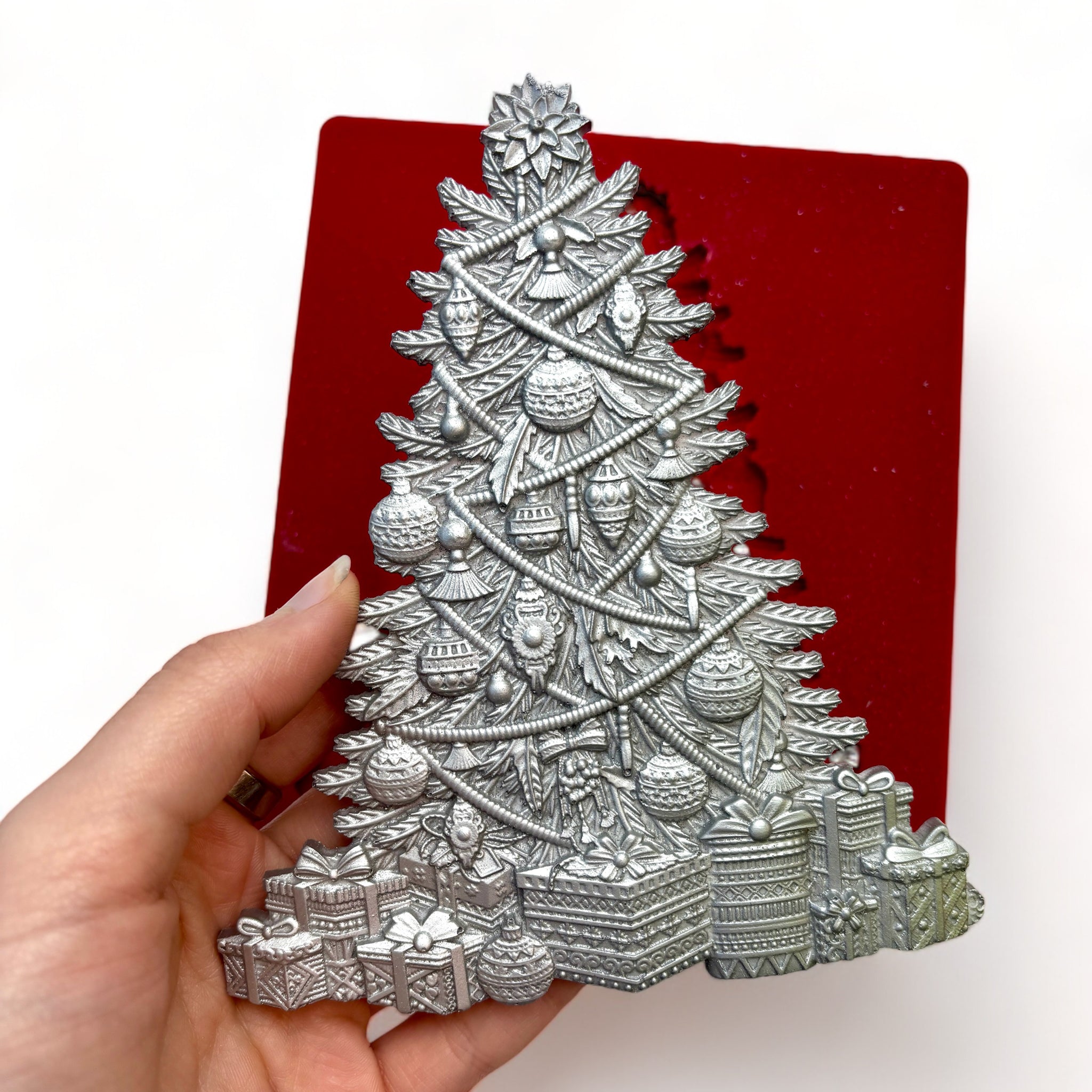 A red silicone mold and silver color casting of Zuri Design's Bohemian Christmas Tree are against a white background. A hand is shown holding the tree casting.