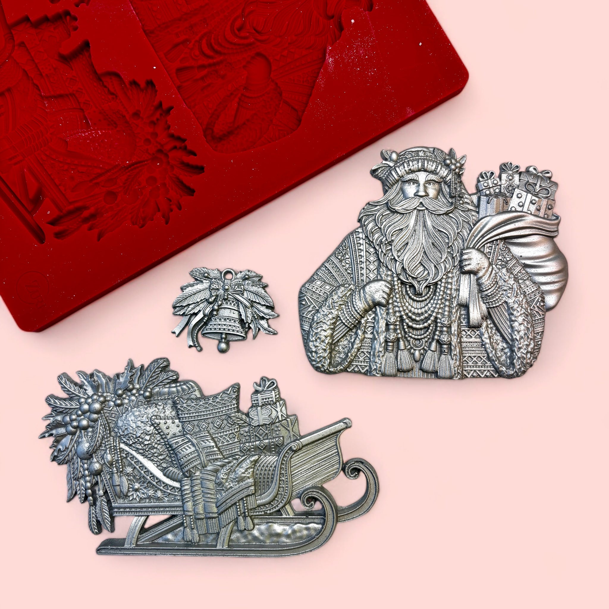 A red silicone mold and silver colored castings of a Bohemian style Santa, his sled, and a bell with foliage are against a light pink background.