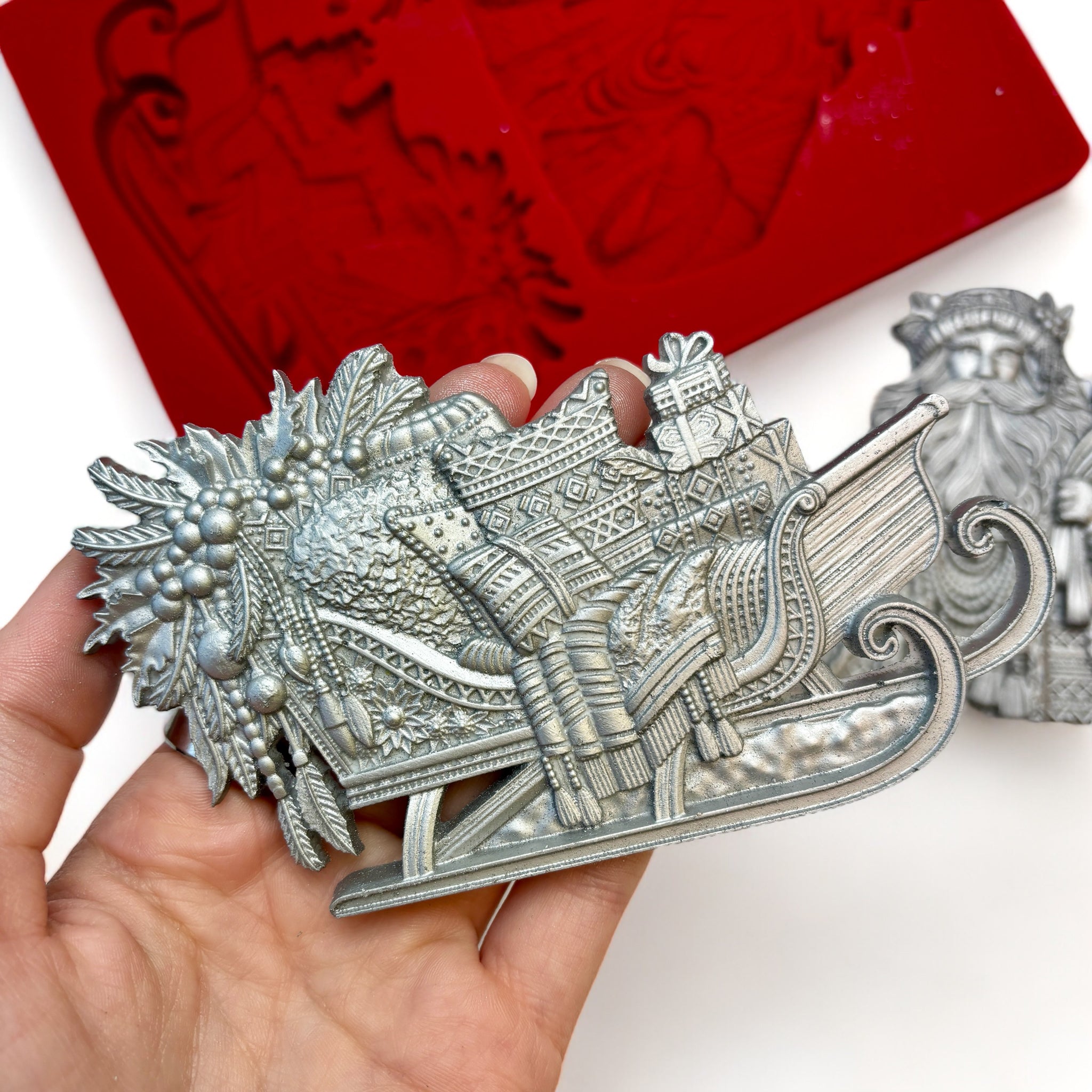 A red silicone mold and silver colored castings of Zuri Design's Bohemian Santa and Sled are against a white background. A hand is shown holding the sled casting.