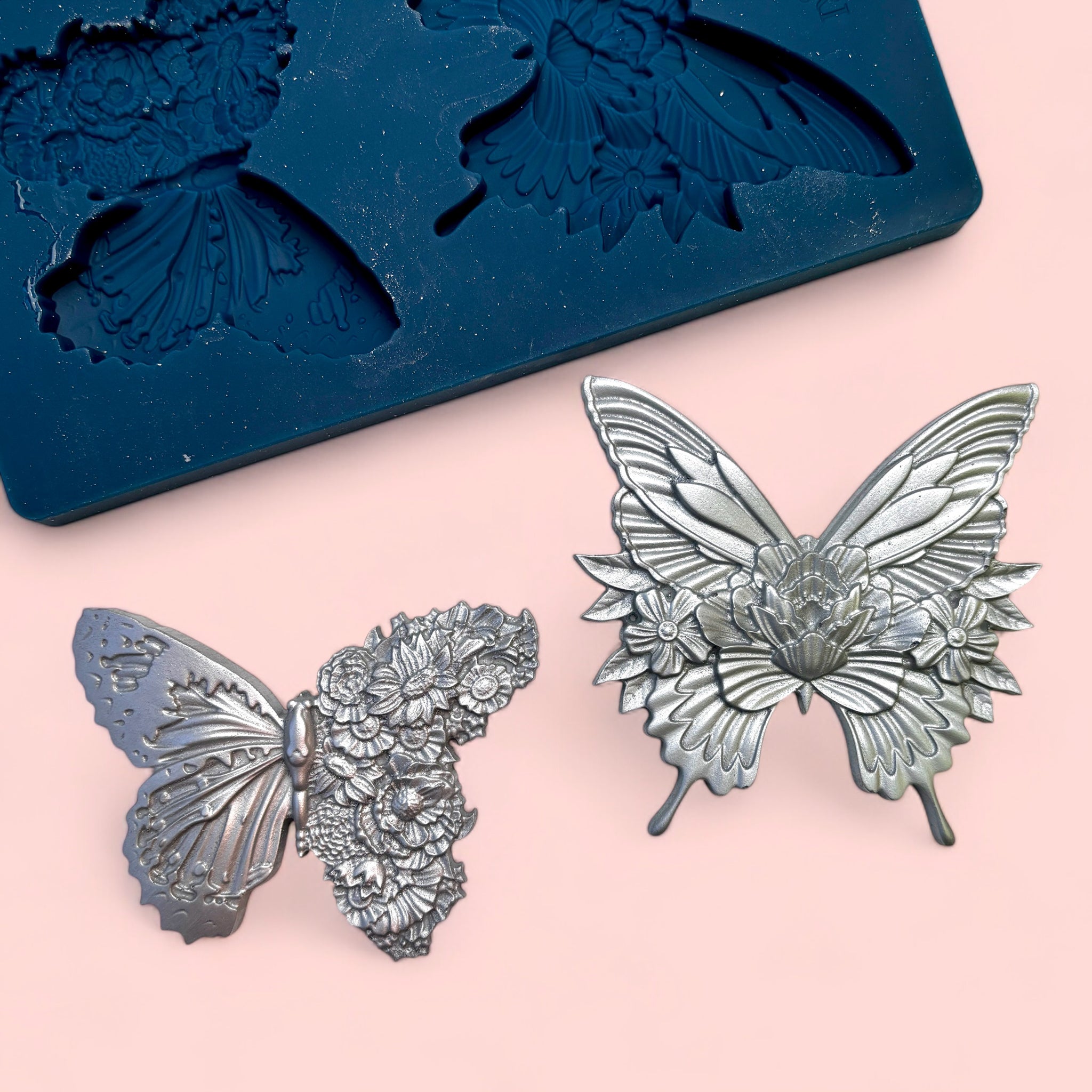 A blue silicone mold and silver colored castings of 2 ornate floral butterflies are against a light pink background.