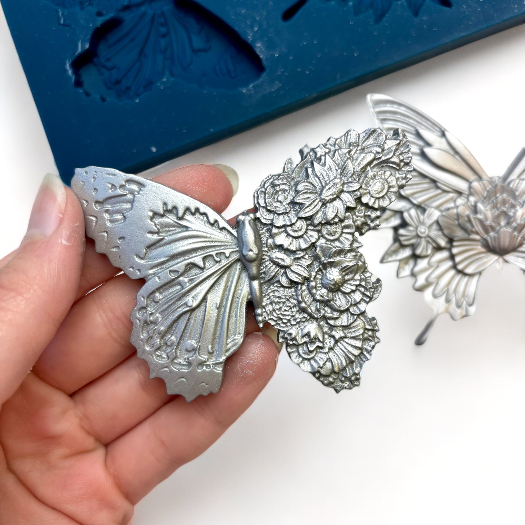 A blue silicone mold and silver colored castings of Zuri Design's Butterfly Dance are against a white background. A hand is shown holding one of the butterfly castings.