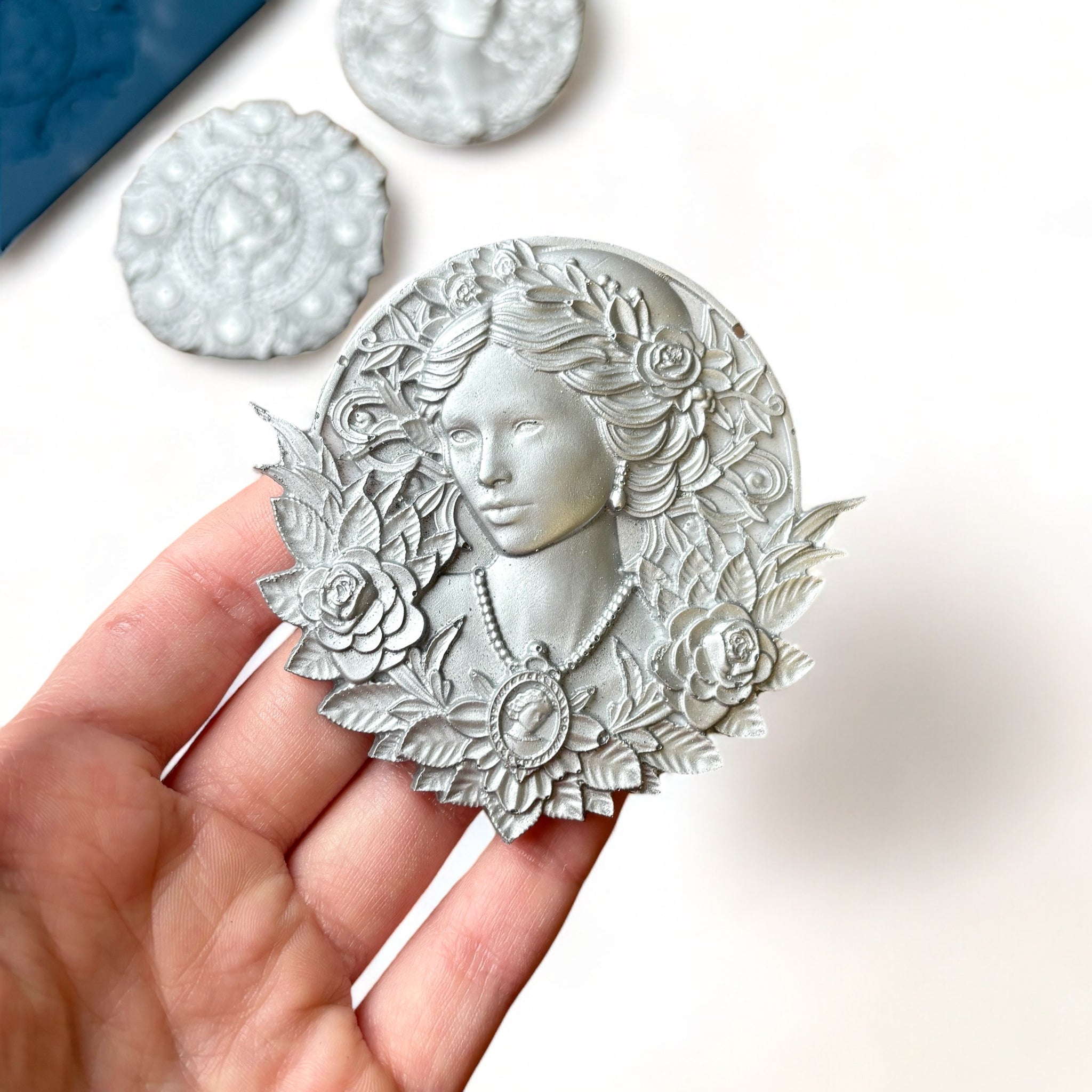 A blue silicone mold and silver colored castings of 3 ornate cameo medallions are against a white background. A hand is shown holding one of the cameo castings.