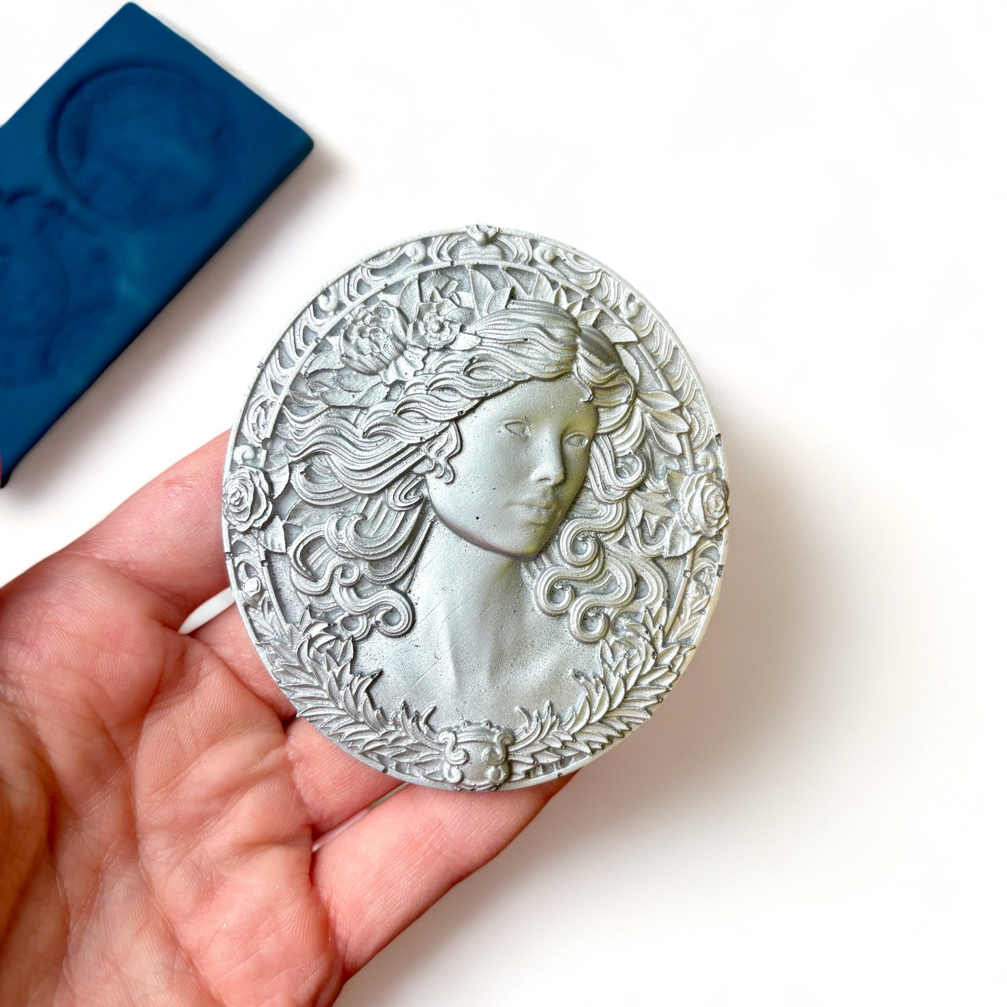 A blue silicone mold and silver colored castings of 3 ornate cameo medallions are against a white background. A hand is shown holding one of the cameo castings.