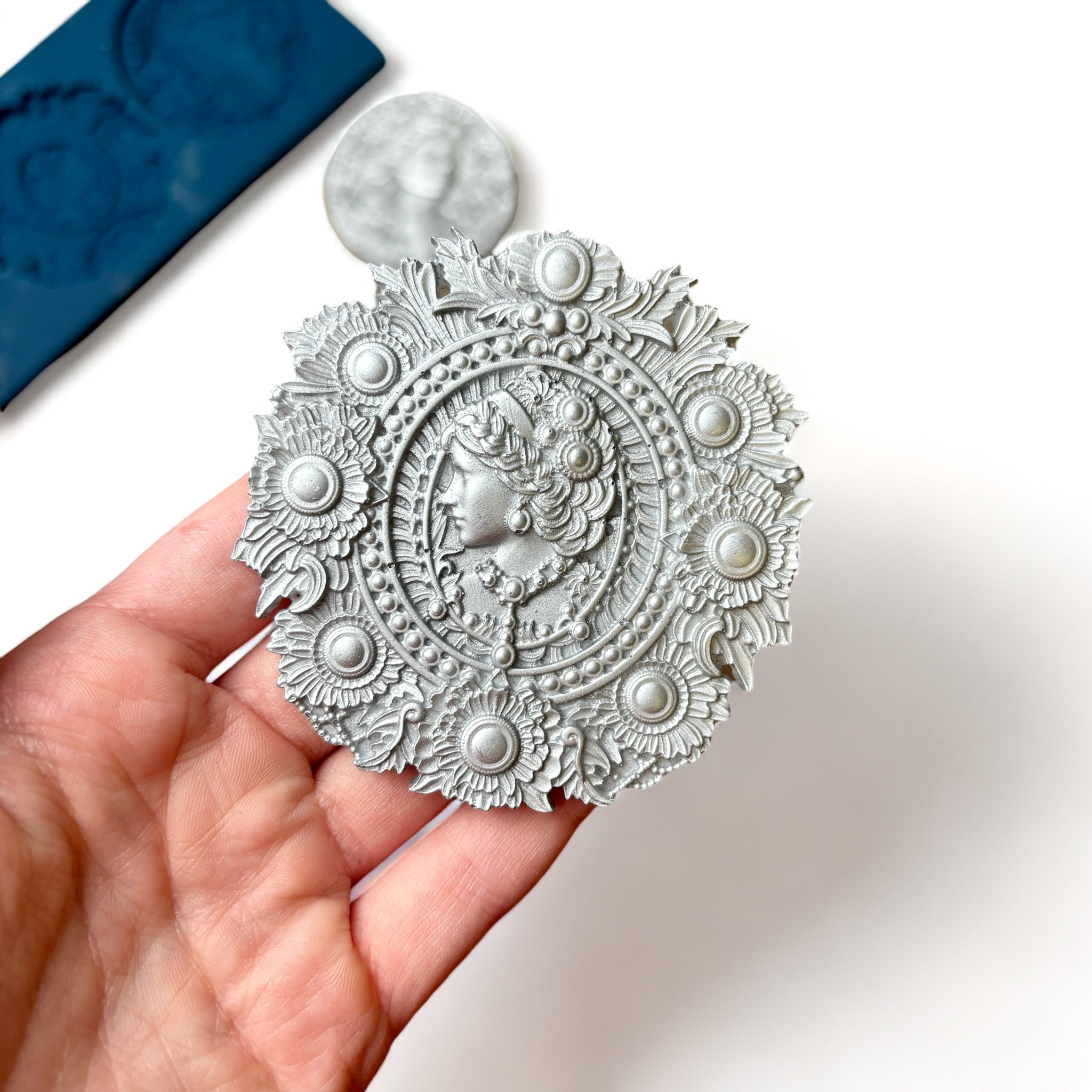 A blue silicone mold and silver colored castings of 3 ornate cameo medallions are against a white background. A hand is shown holding one of the cameo castings.