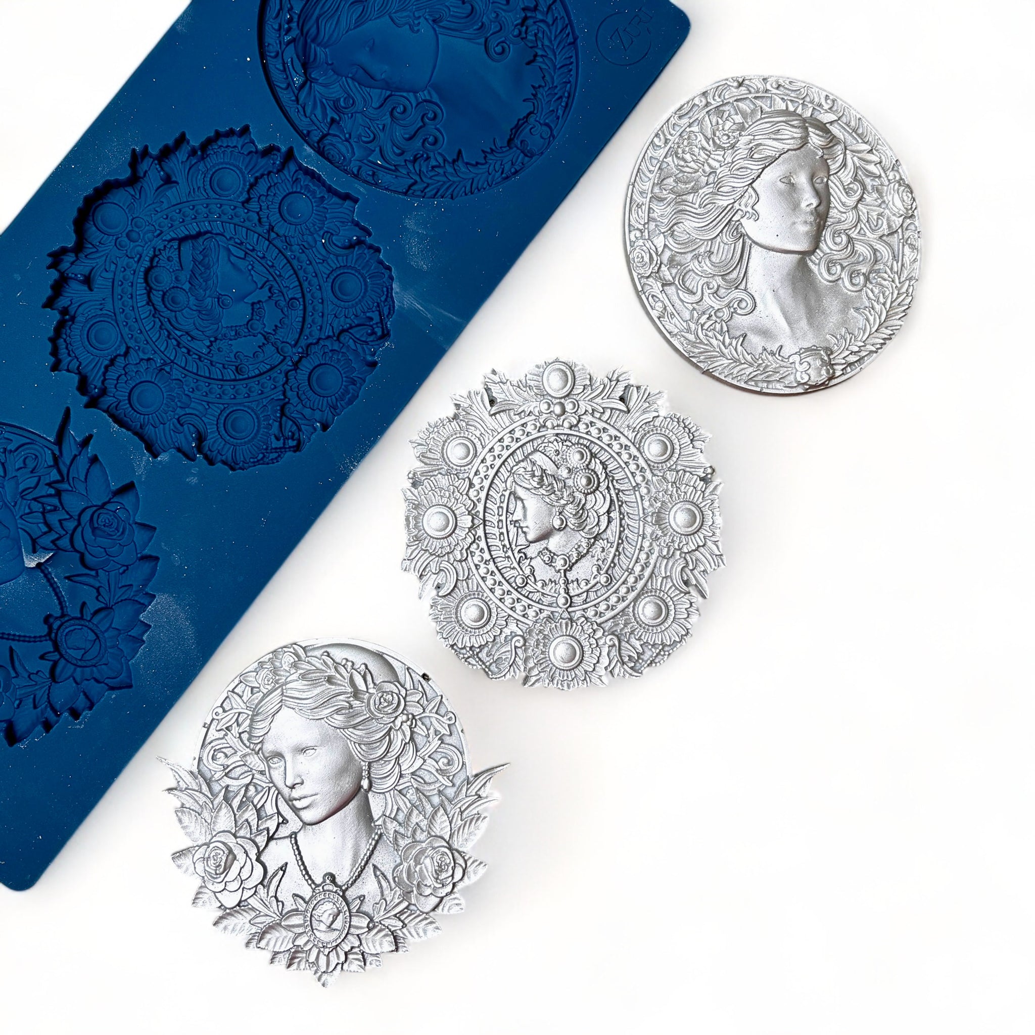 A blue silicone mold and silver colored castings of 3 ornate cameo medallions are against a white background.