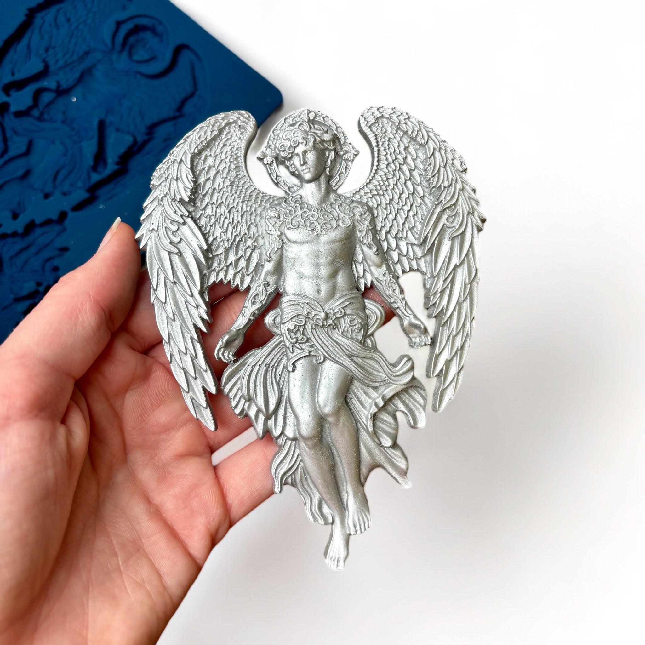 A blue silicone mold featuring 1 large, and 2 small celestial angels with large wings is against a white background. A hand is shown holding a silver colored casting of the large angel.