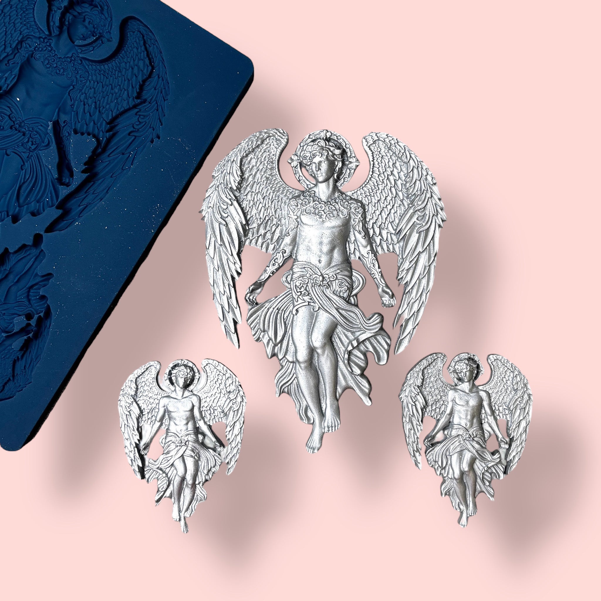 A blue silicone mold and silver colored castings of 1 large, and 2 small celestial angels with large wings are against a light pink background.