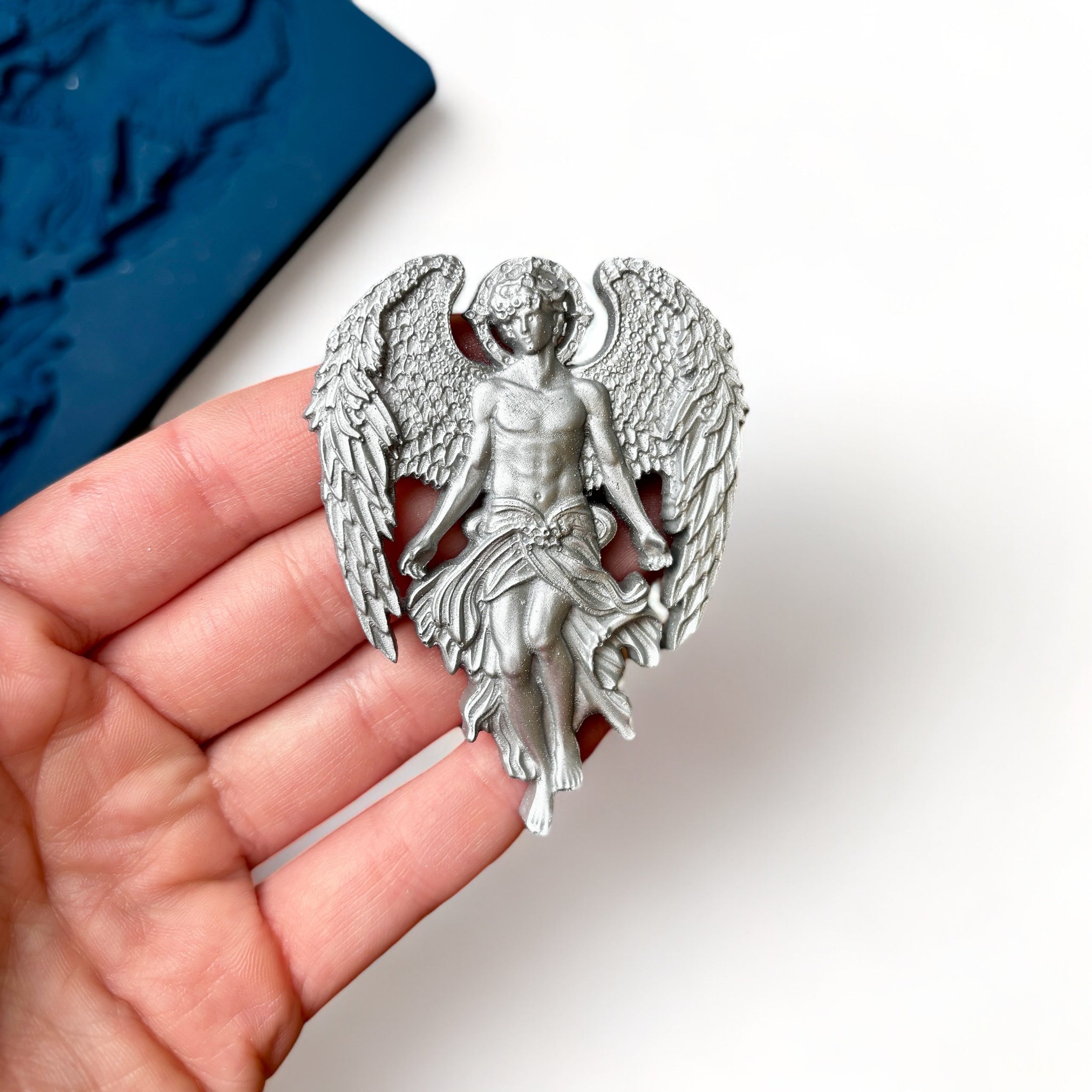 A blue silicone mold featuring 1 large, and 2 small celestial angels with large wings is against a white background. A hand is shown holding a silver colored casting of one of the small angels.